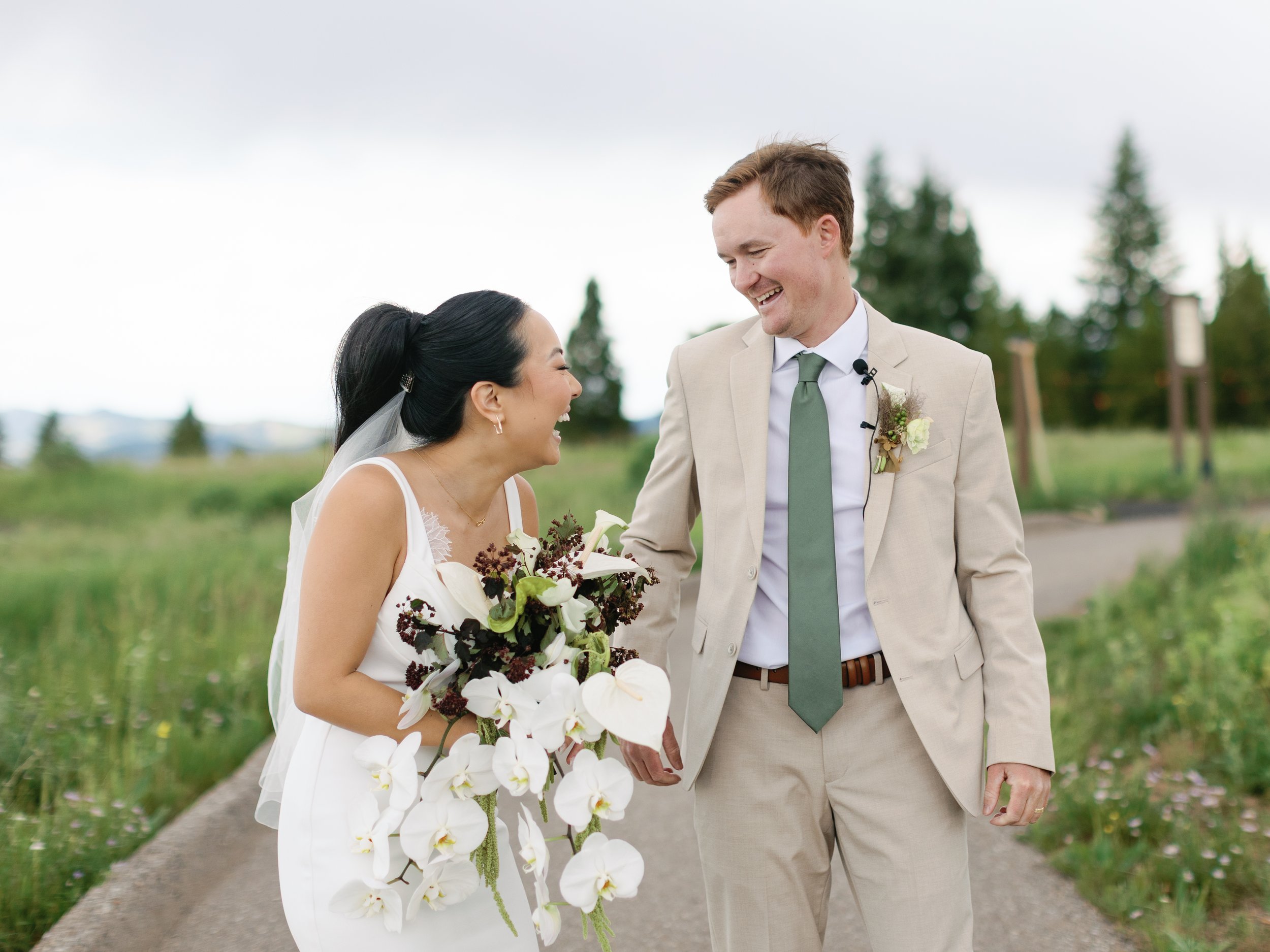 A bride and groom smiling and holding hands outdoors on their wedding day, with trees and a cloudy sky in the background.