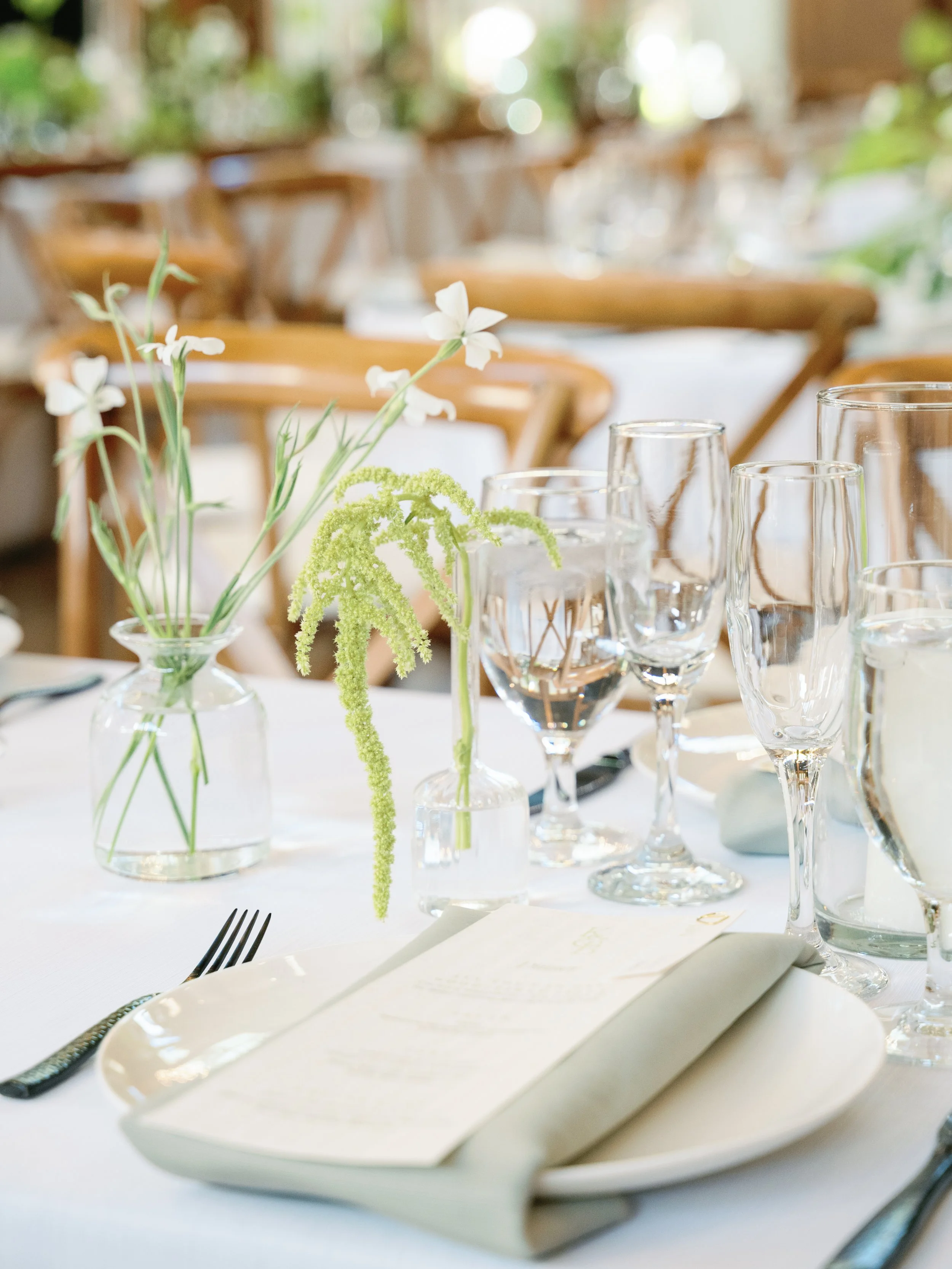 Elegant table setting with floral centerpieces, glassware, and place settings at a well-lit dining area with wooden chairs.