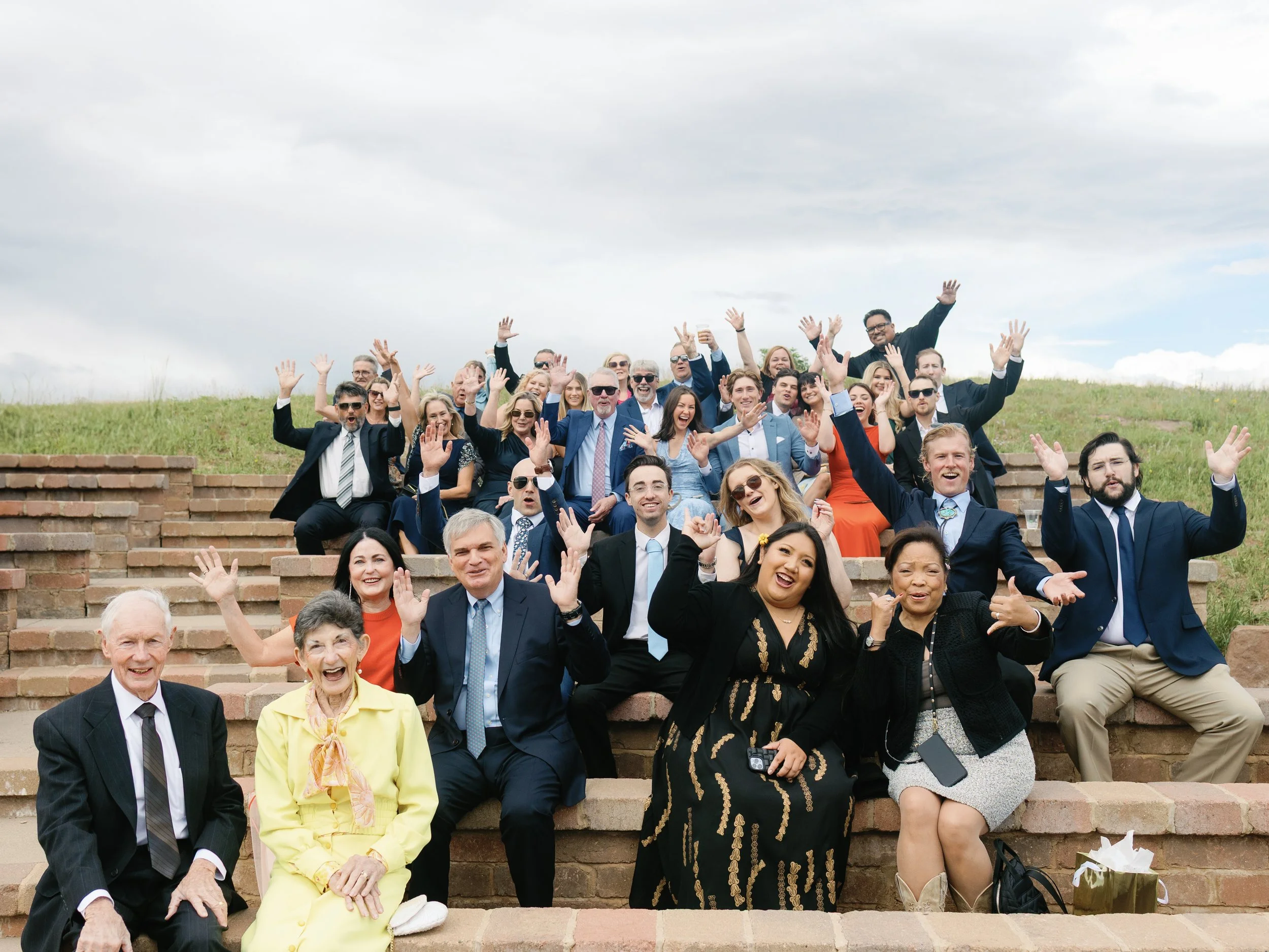 A large group of people dressed in formal attire sitting on outdoor brick steps, smiling, and raising their hands, with green grass and cloudy sky in the background.