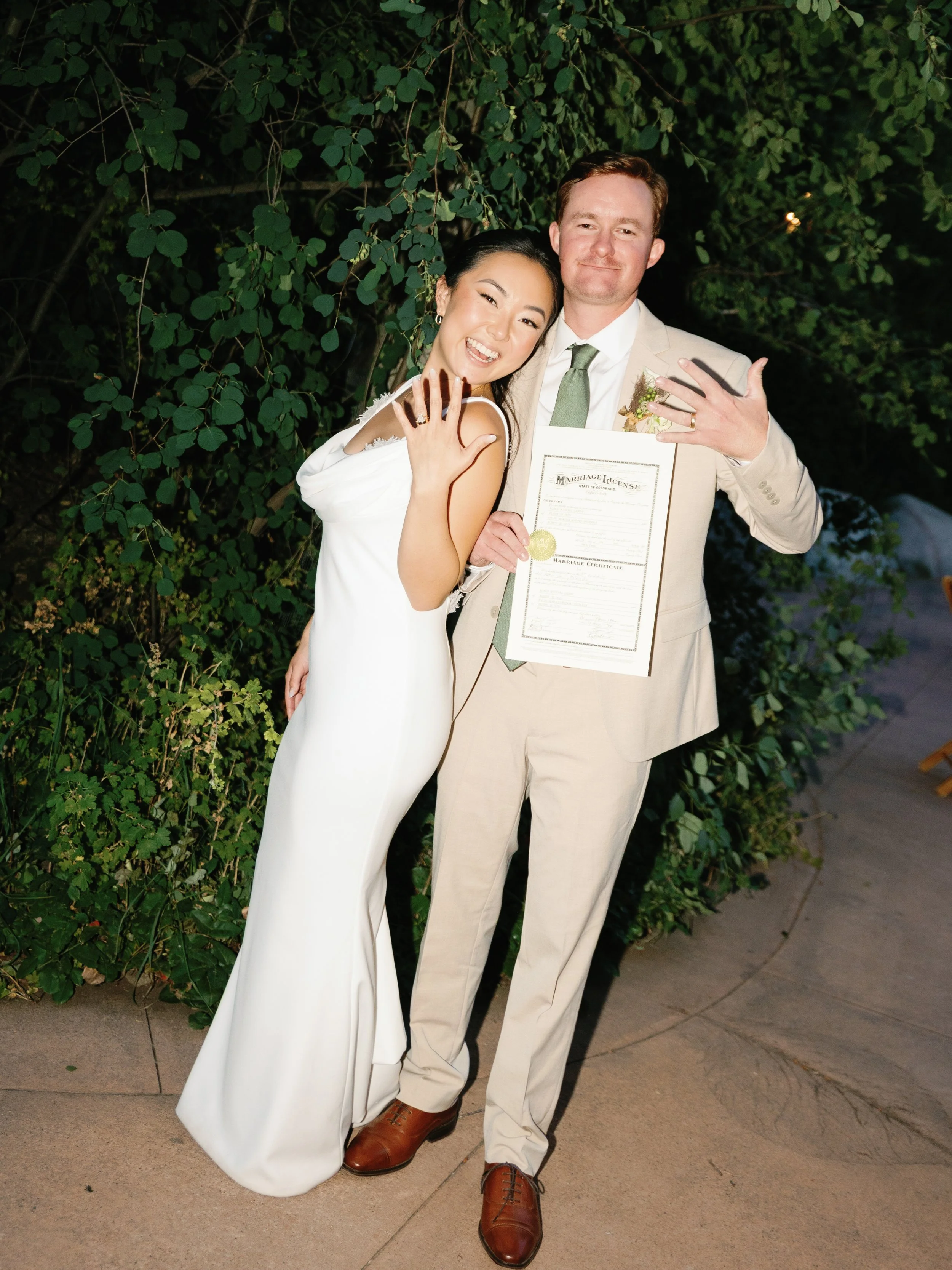 A newly married couple celebrating outdoors, showing off their wedding rings and certificate.