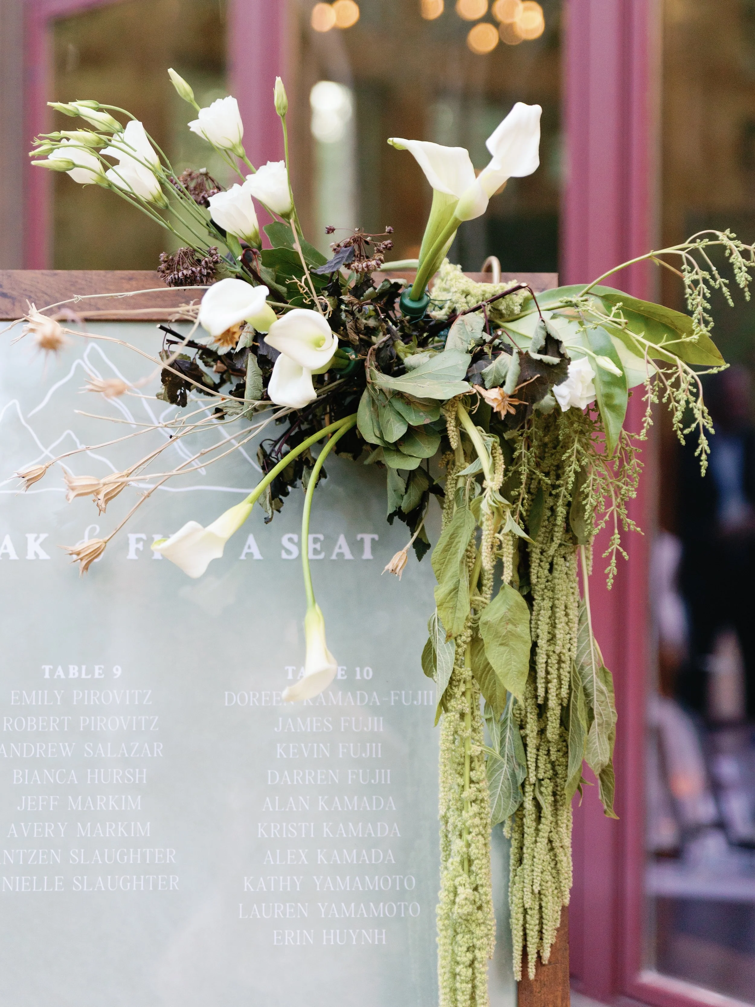A floral arrangement with white flowers, green leaves, and hanging green amaranthus, placed next to a glass sign with text on it.