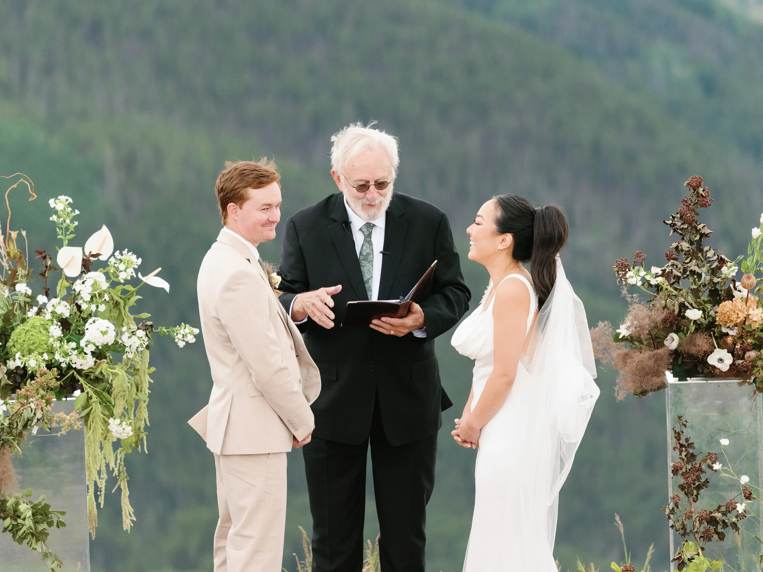 A wedding ceremony outdoors with a groom in a beige suit and a bride in a white dress standing facing each other, with an officiant in a black suit holding a book and smiling. The background shows a scenic mountain landscape, and floral arrangements 