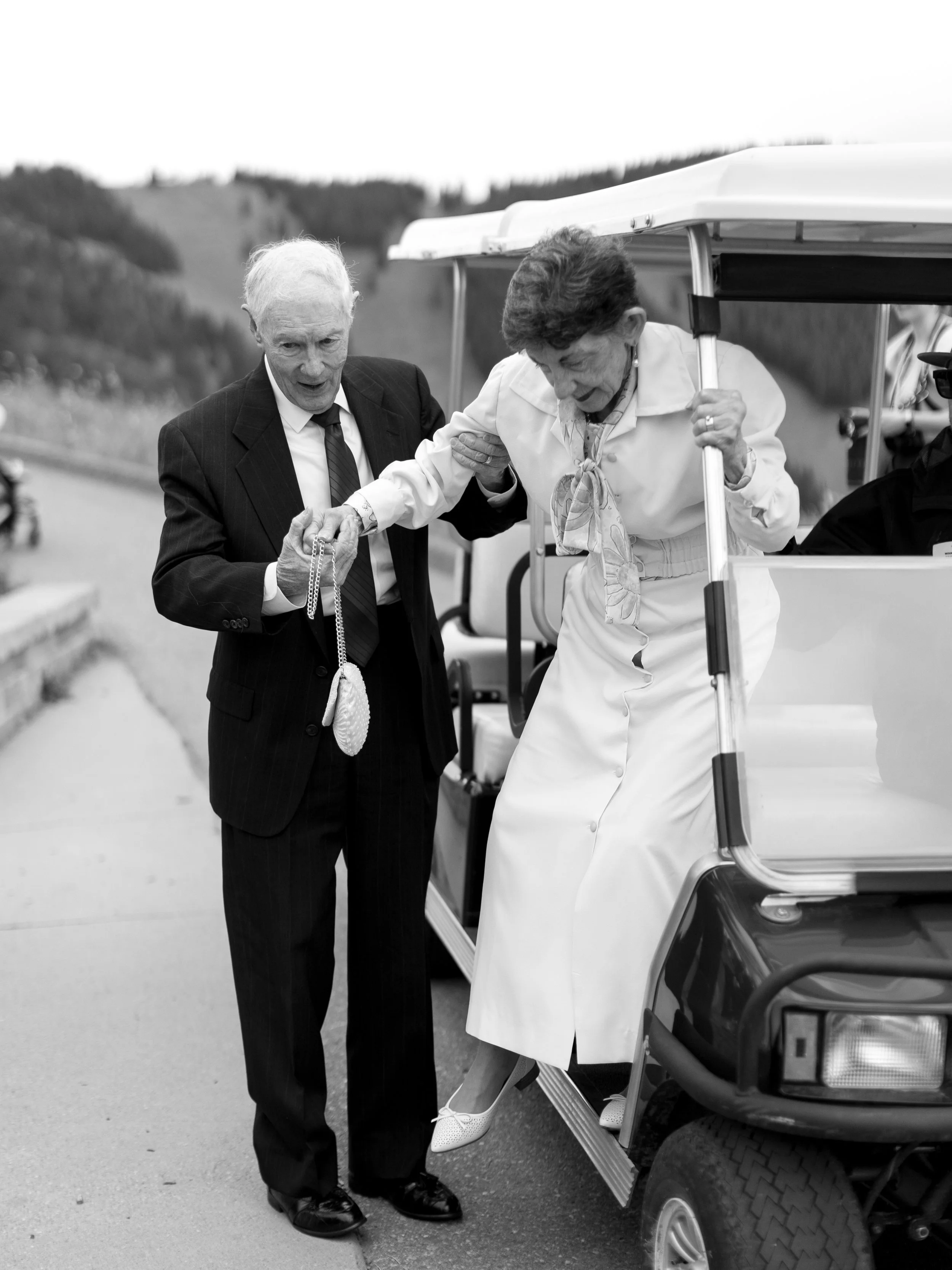 An elderly man in a suit helps an elderly woman get onto a golf cart, holding a purse with a chain. The woman is holding onto a pole of the golf cart and stepping onto it, dressed in a white dress and shoes. The scene takes place outdoors with hills 