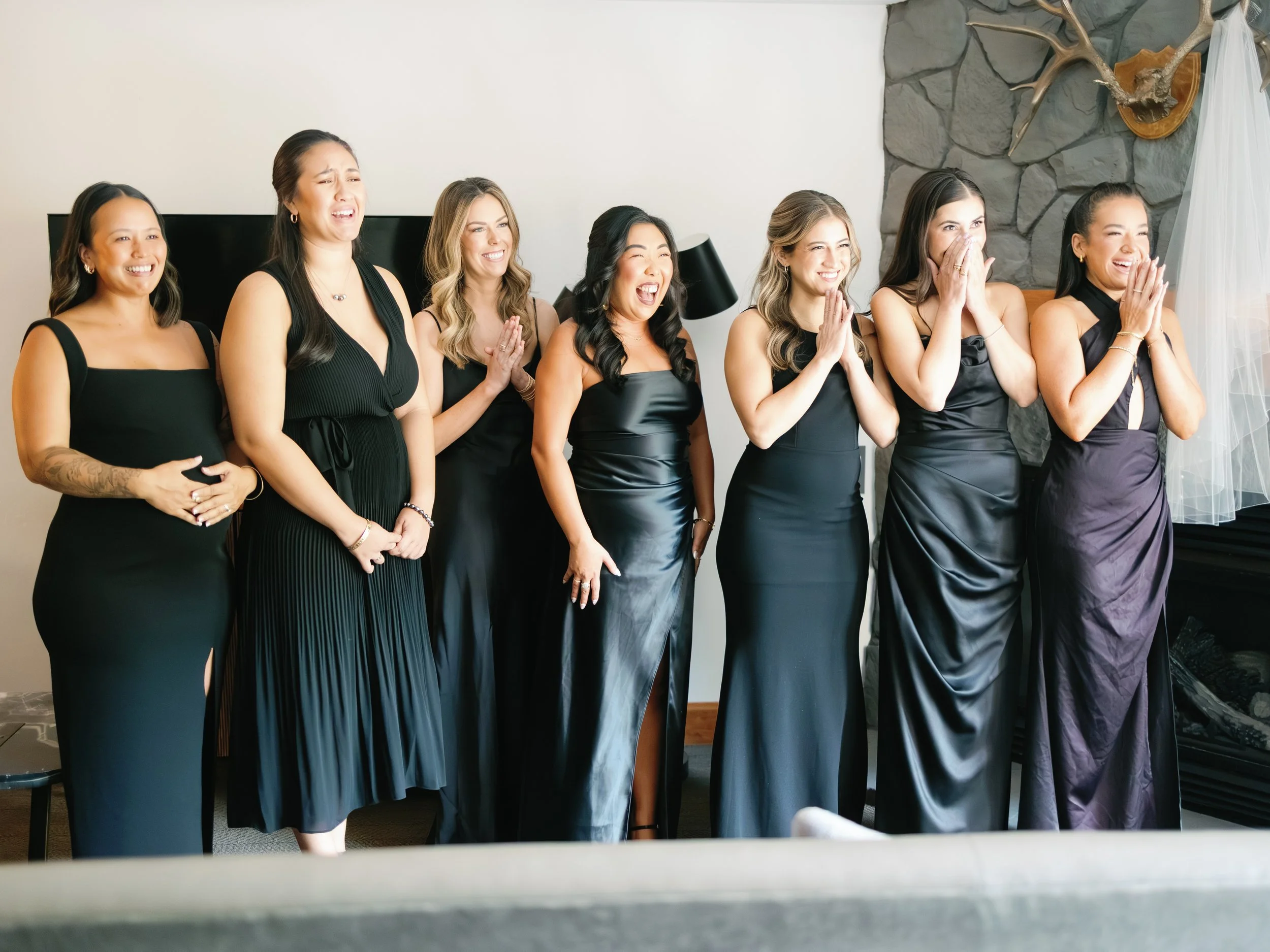 A group of seven women dressed in black dresses, laughing and looking towards the right side of the image, standing indoors in front of a white wall with a stone accent wall and deer antlers mounted on it.
