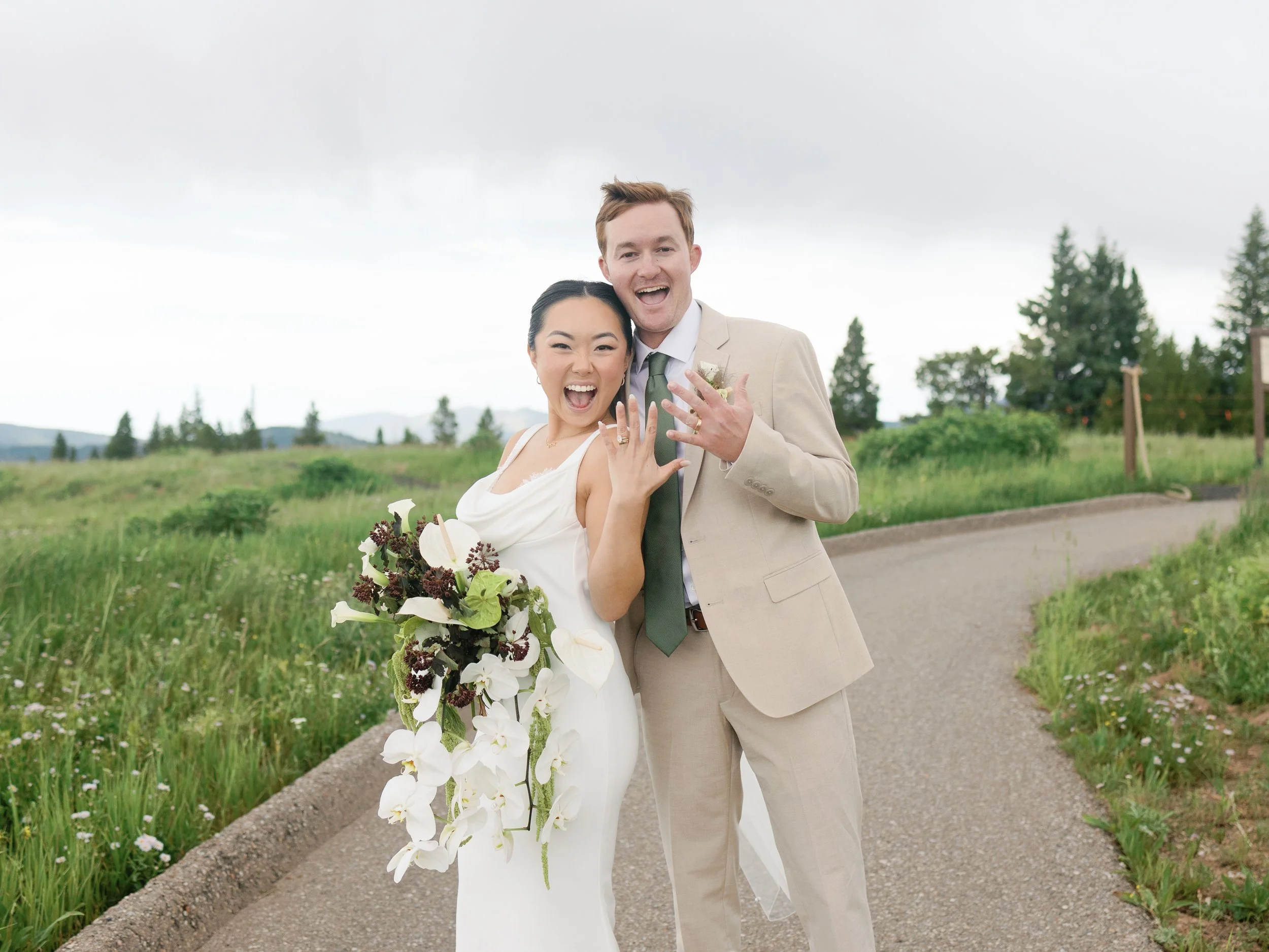 A happy newlywed couple posing outdoors on a winding pathway in a green field with trees in the background, showing off their wedding rings.