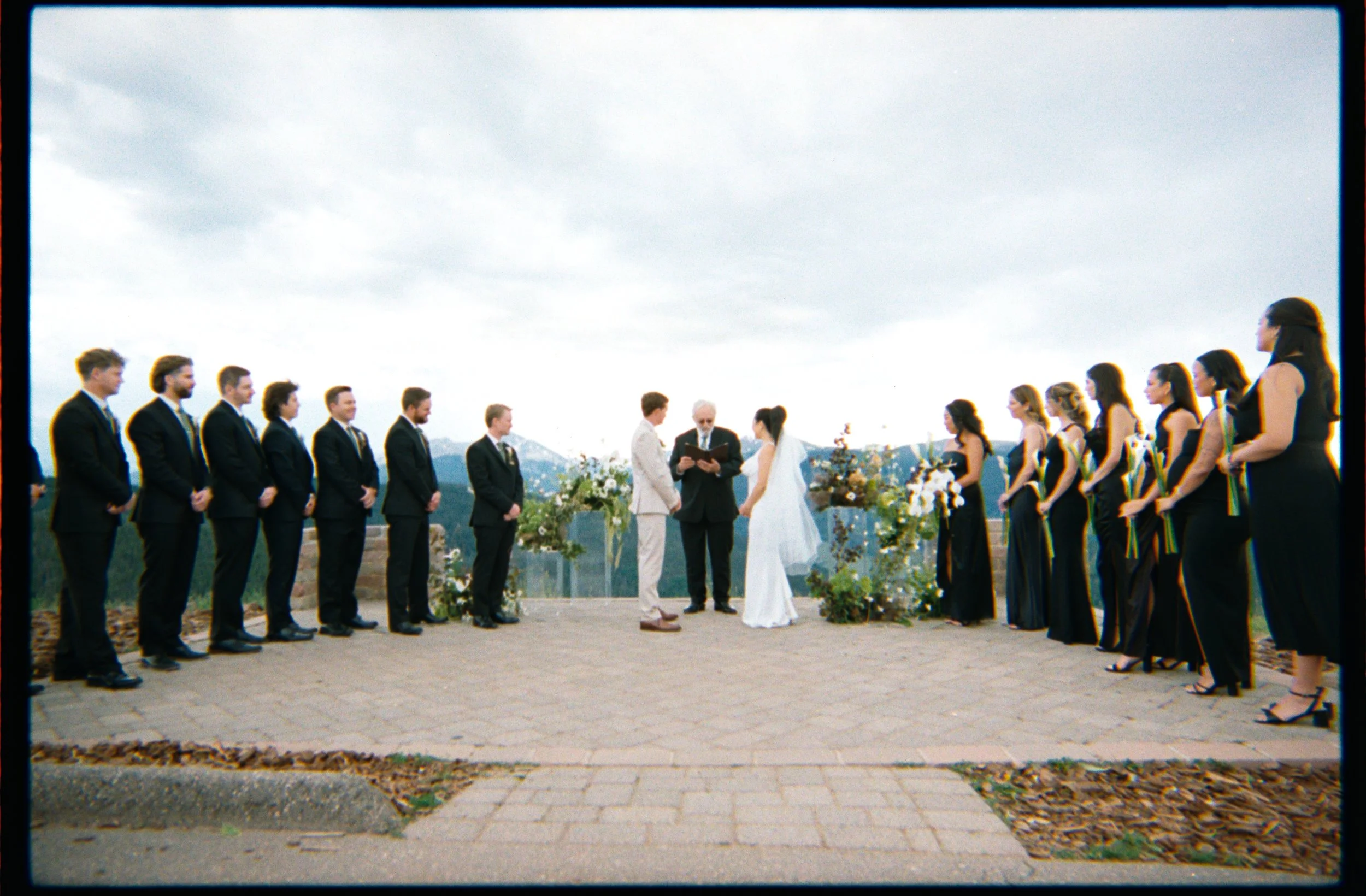A wedding ceremony outdoors with the bride and groom standing before an officiant, surrounded by their wedding party and guests, with scenic mountains in the background.