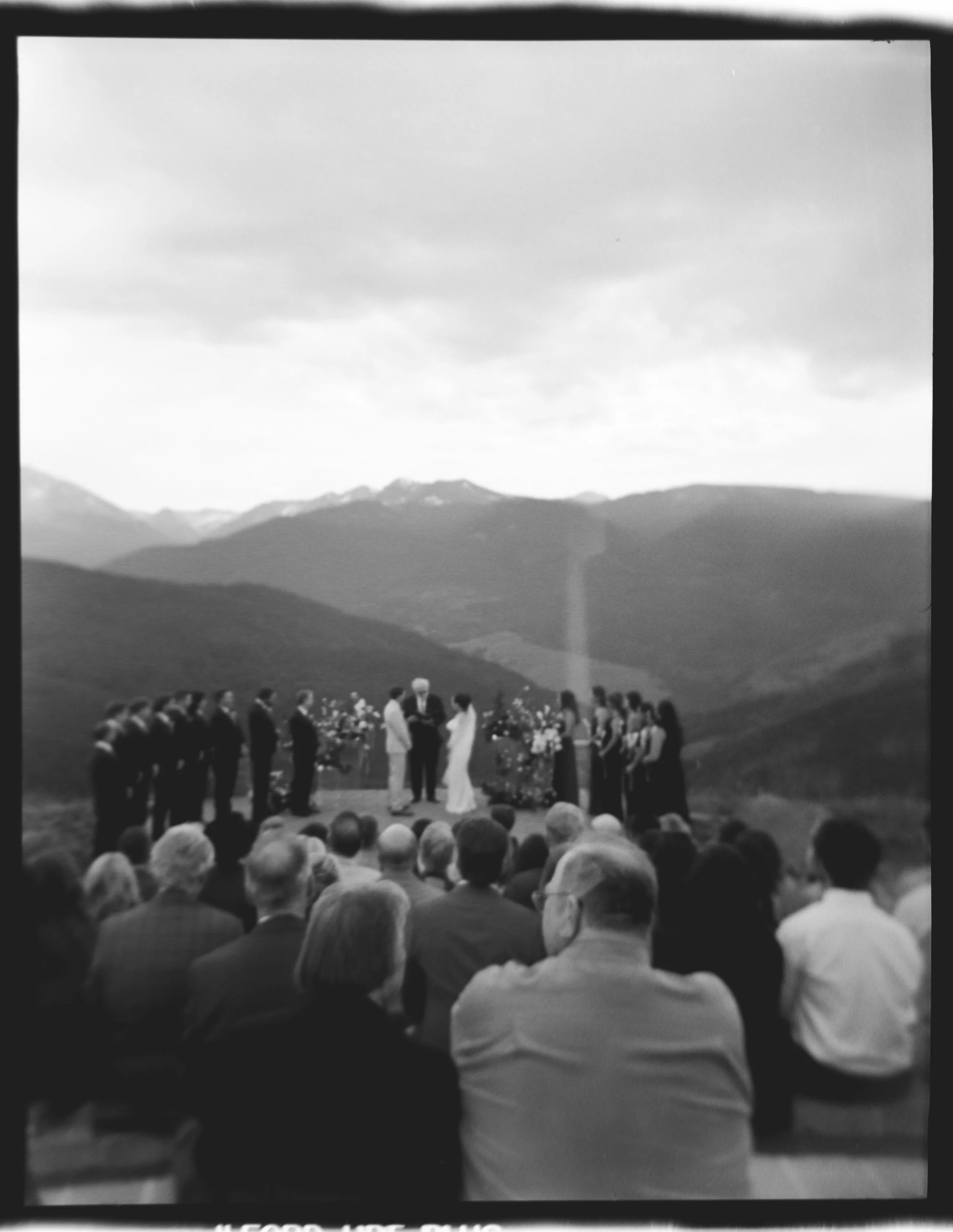 A black-and-white outdoor wedding ceremony with mountains in the background, where a couple is exchanging vows, surrounded by guests seated on benches.