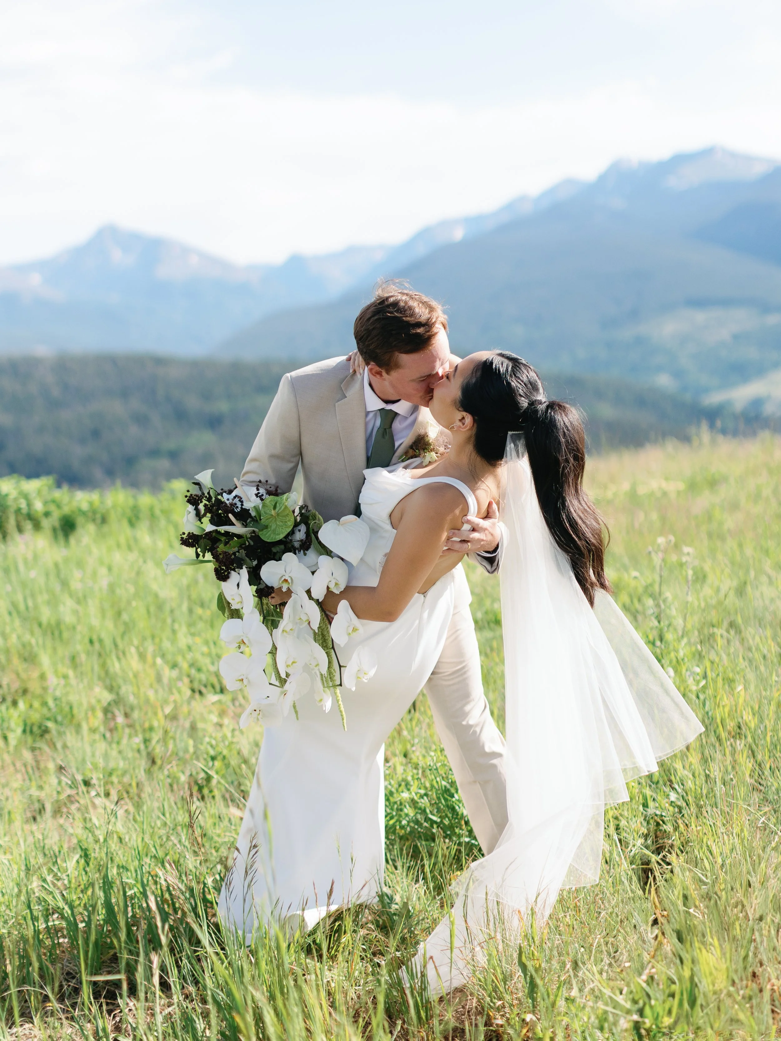 A bride and groom sharing a kiss outdoors on a grassy field with mountains in the background, the bride holding a bouquet of white and dark flowers.