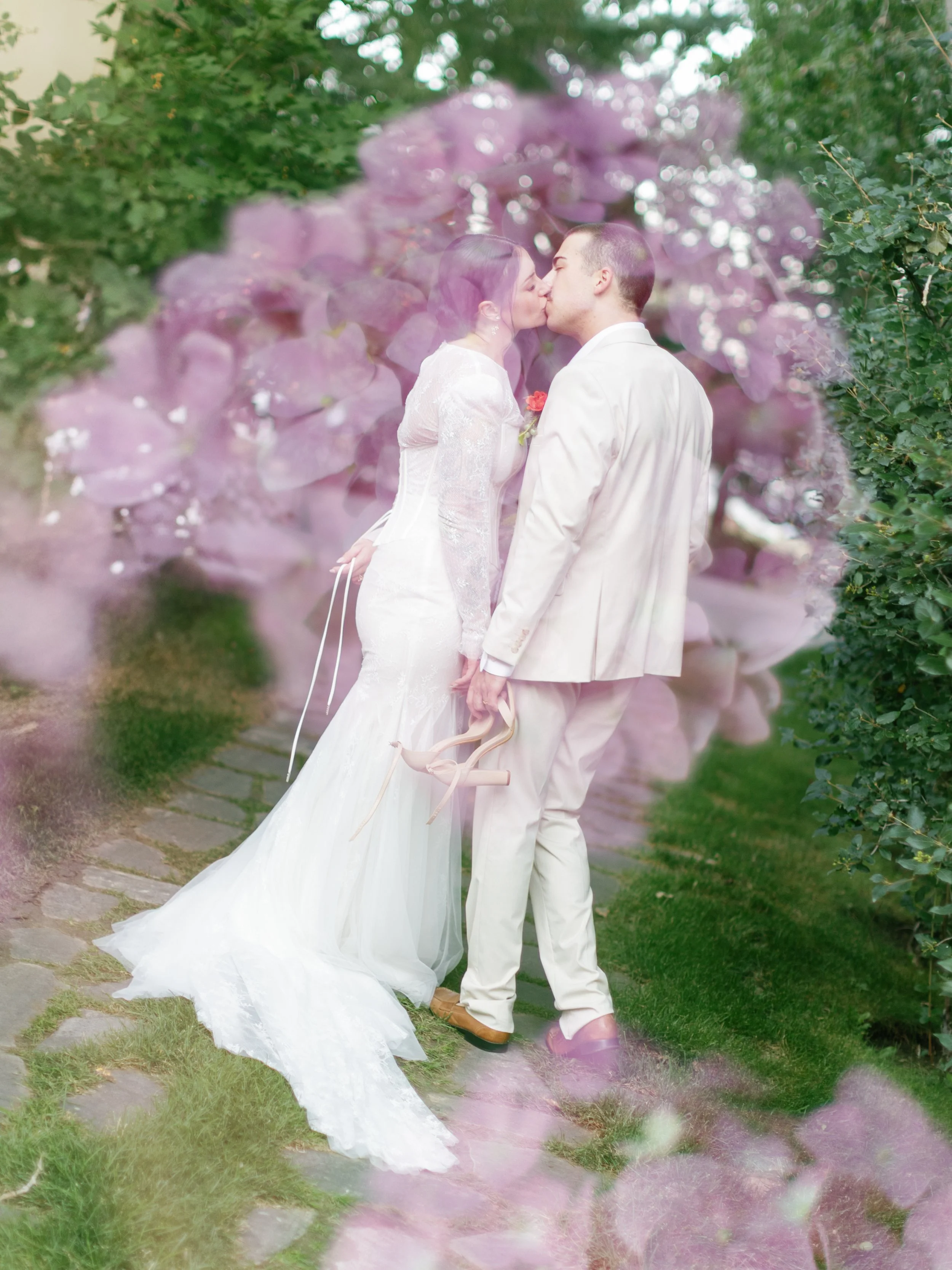 A bride and groom share a kiss outdoors, surrounded by pink flowers and green foliage, with the bride holding a pair of high heels.