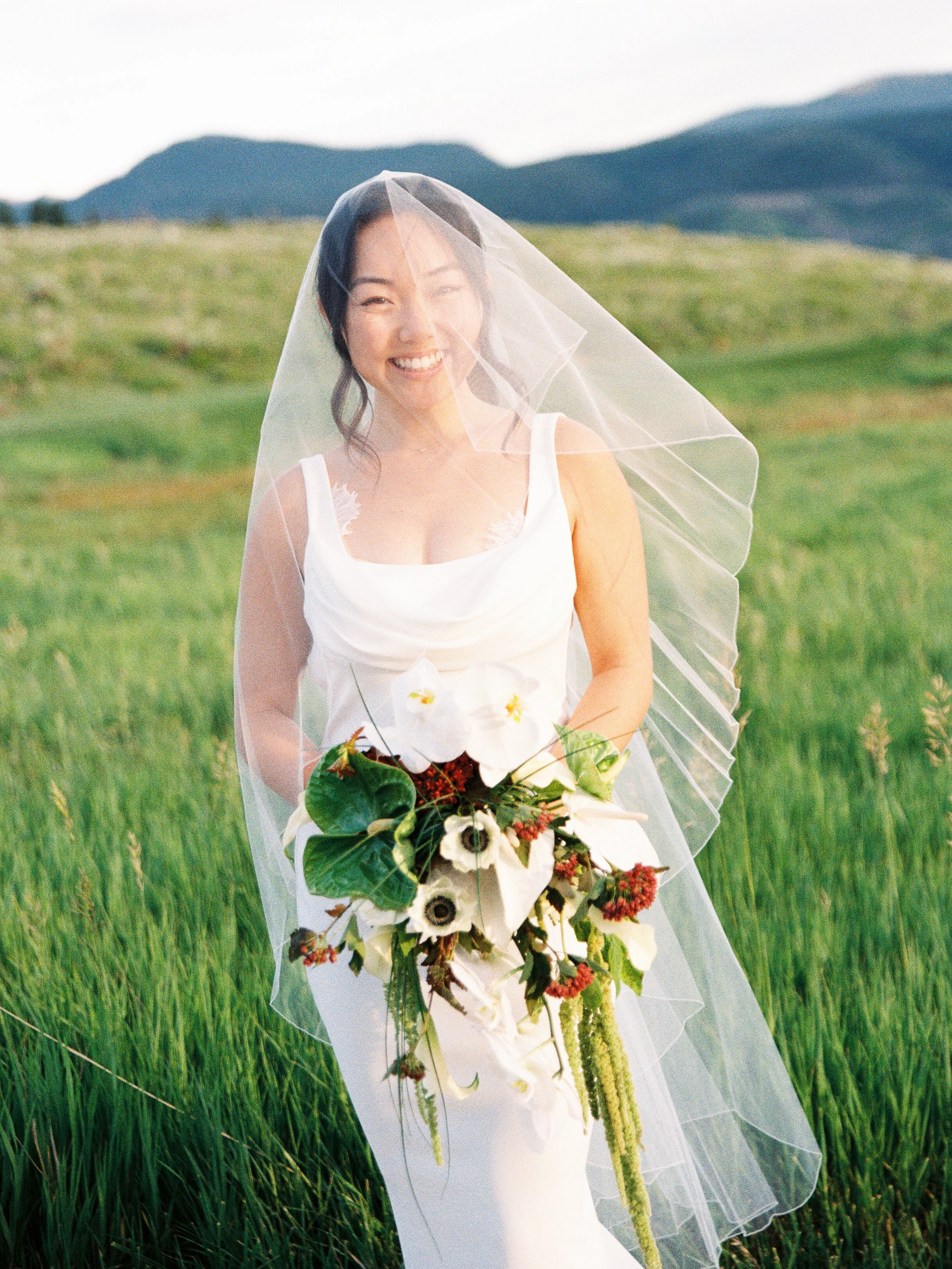 A smiling bride in a white wedding dress holding a bouquet of flowers, standing in a grassy field with mountains in the background.