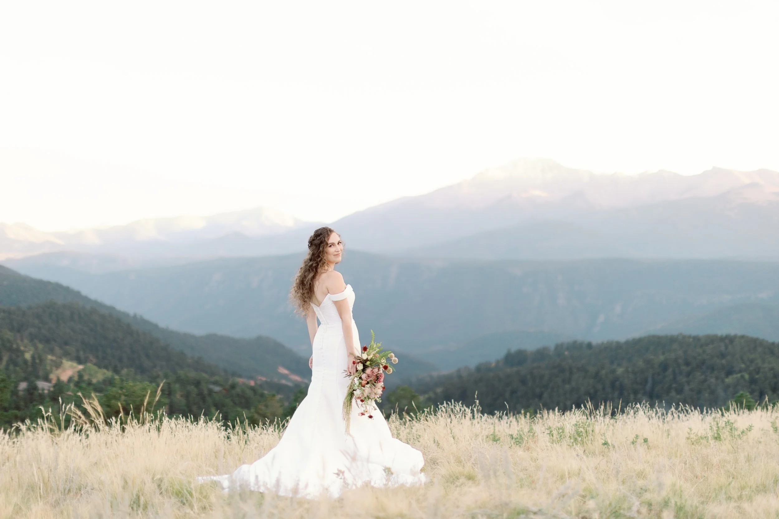 A bride in a white wedding dress standing in a grassy field with mountains in the background, holding a bouquet of flowers.
