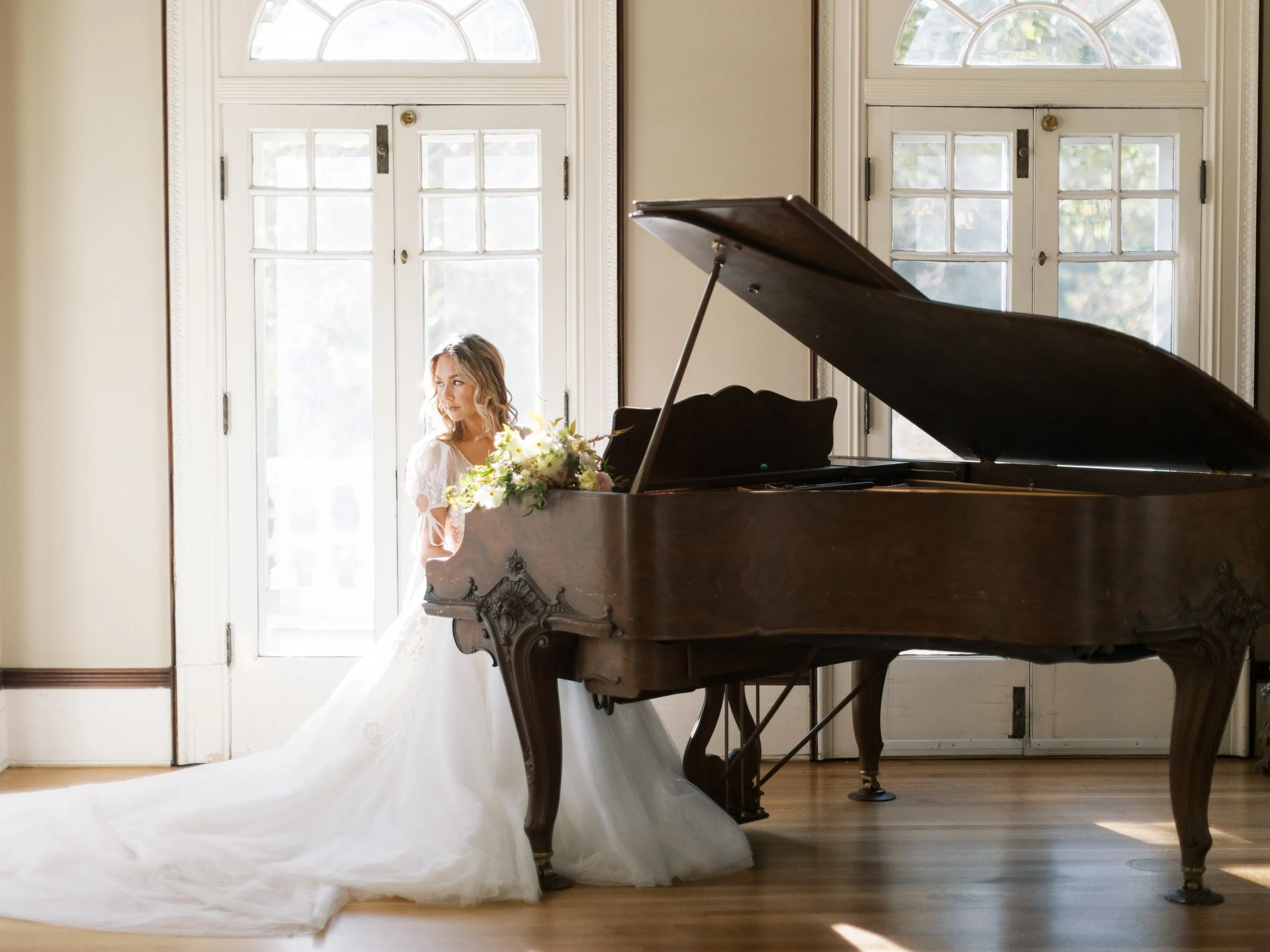 A woman in a wedding dress standing beside a grand piano in a well-lit room with large windows.