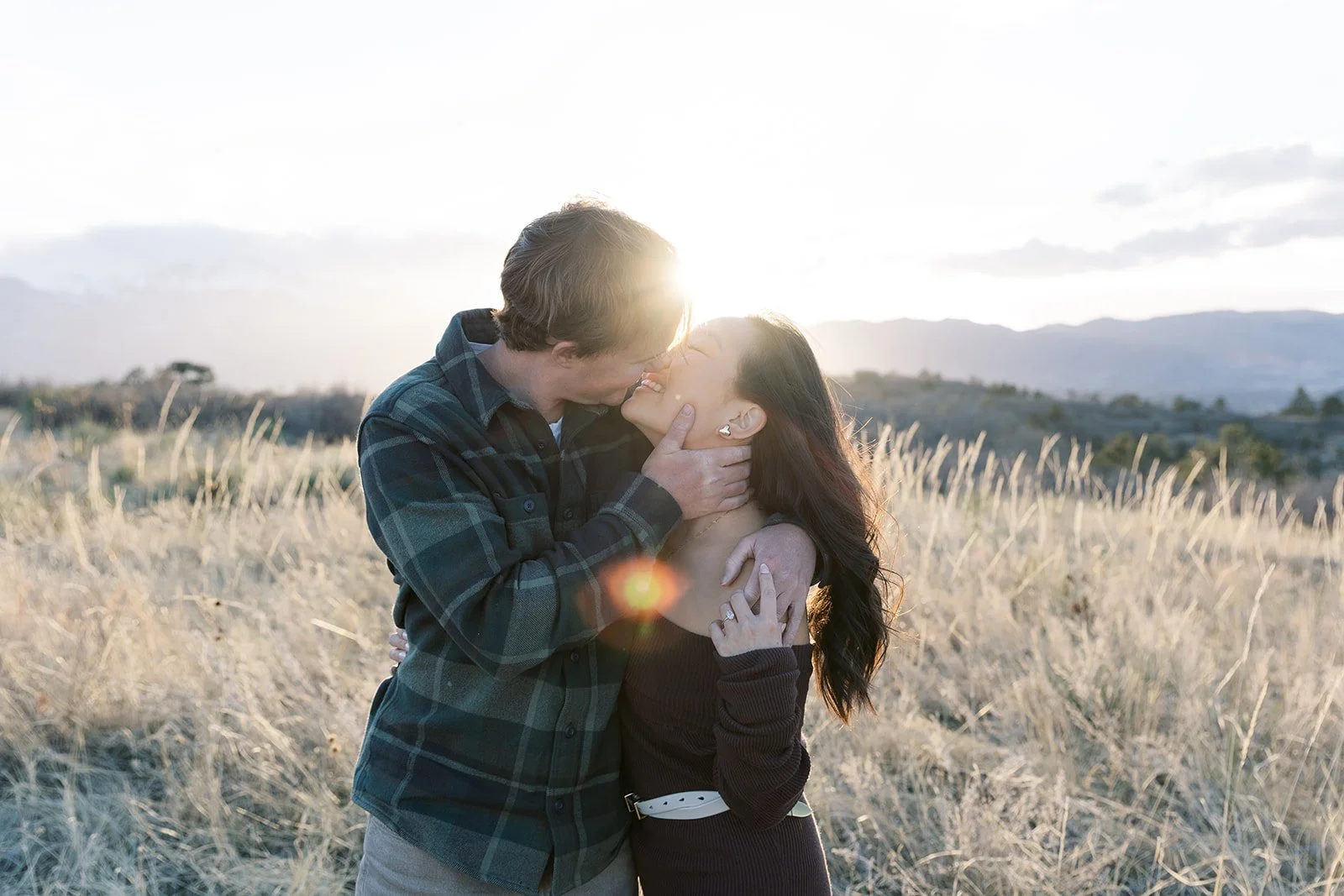 A couple sharing a kiss in a field with mountains in the background at sunset.