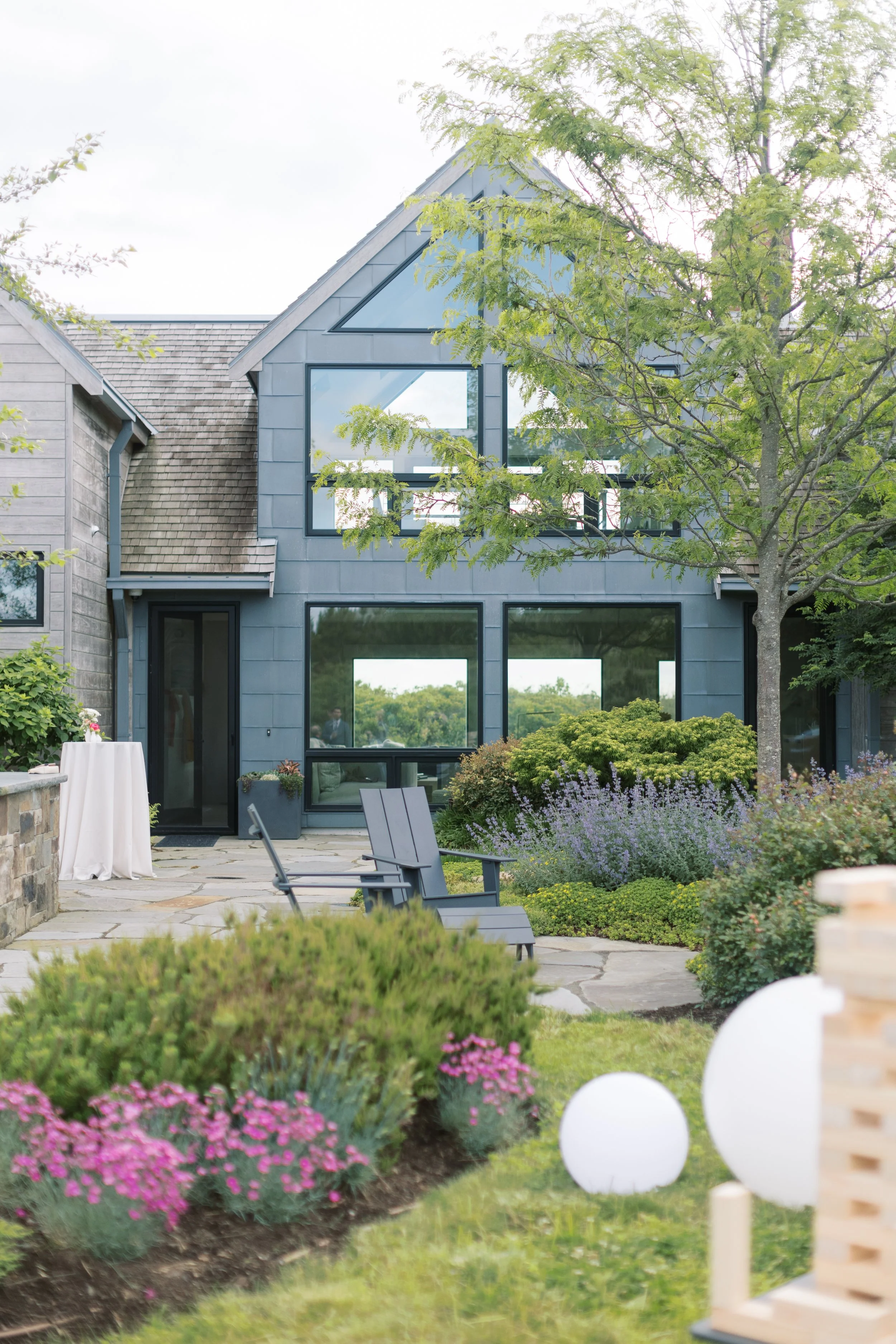 Modern house with large windows surrounded by a landscaped garden with bushes, flowers, and trees, and outdoor seating.