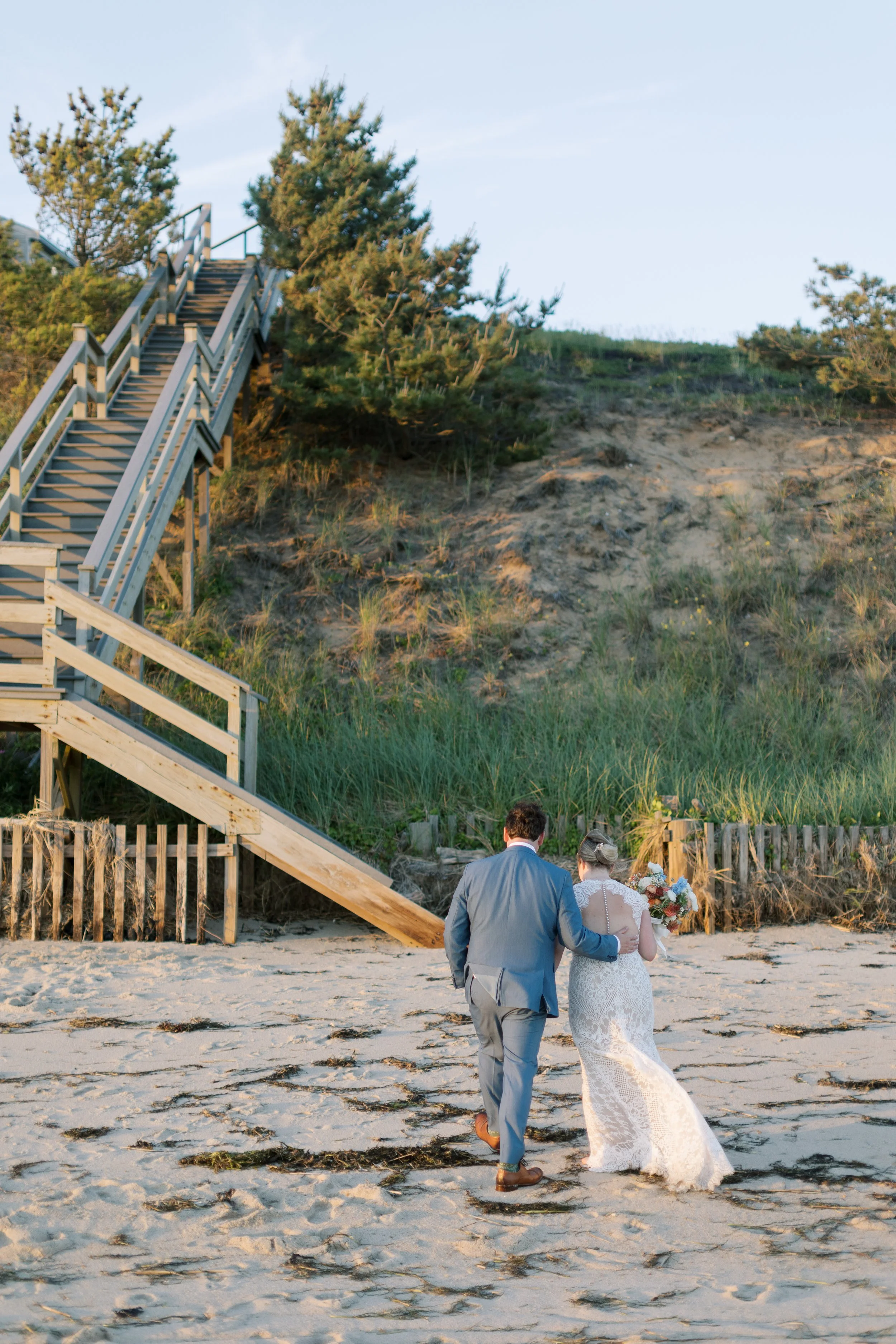 A couple dressed in wedding attire walking on the beach, with a staircase leading up a sand dune in the background.
