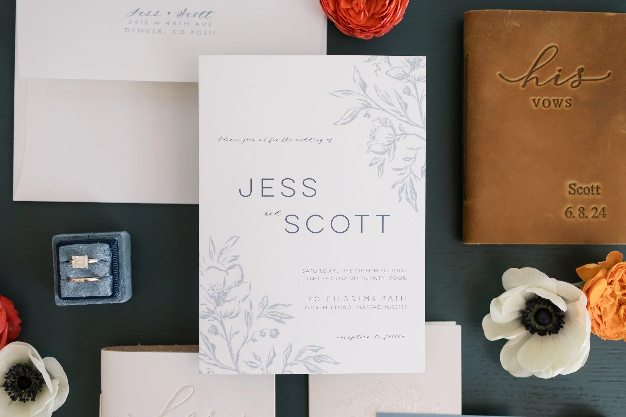 Wedding invitation suite with floral design, ring box, and floral embellishments on dark table. Includes invitation, RSVP card, and a leather item engraved with wedding details.