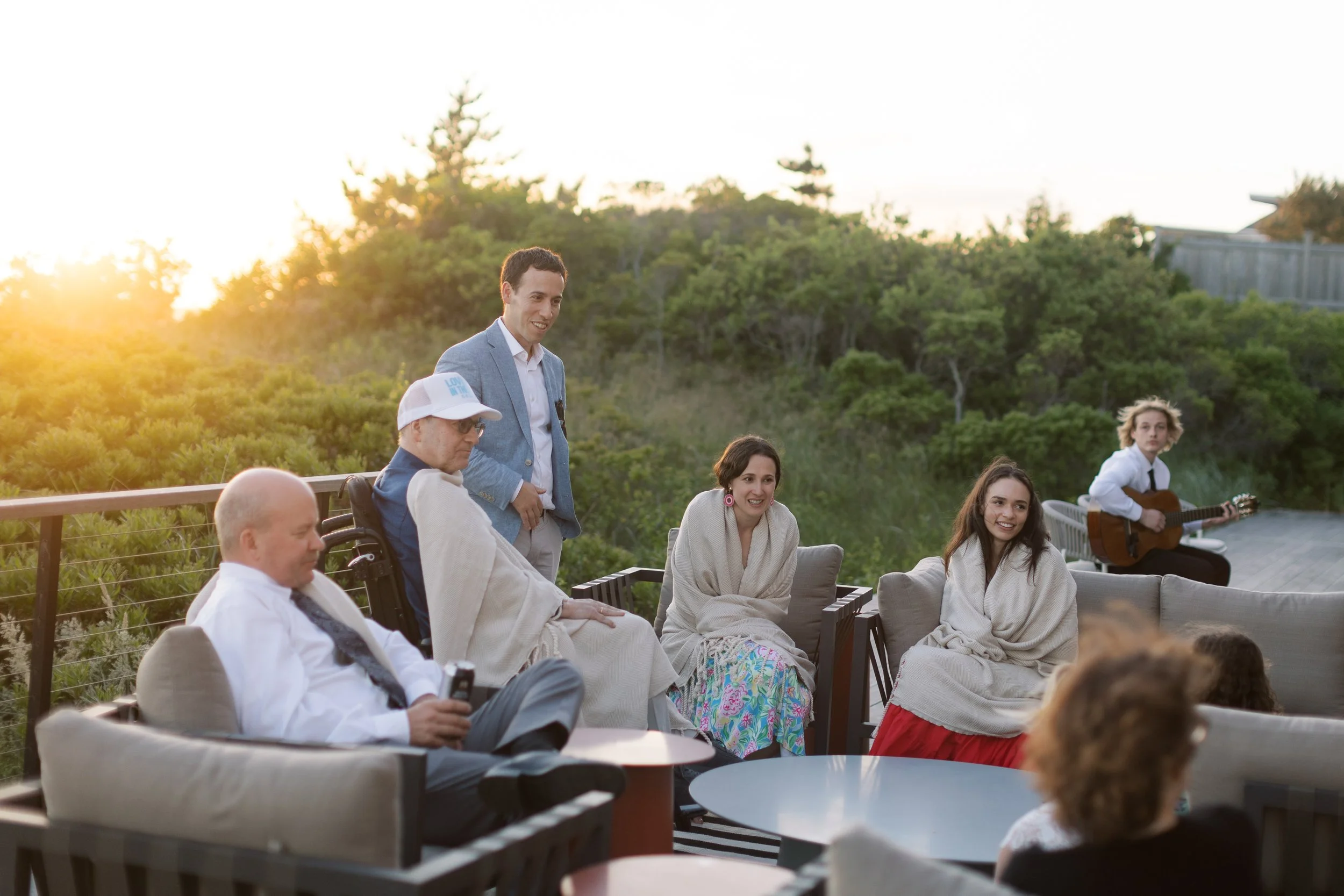 People gathered outdoors on a terrace during sunset, some sitting and one playing guitar.