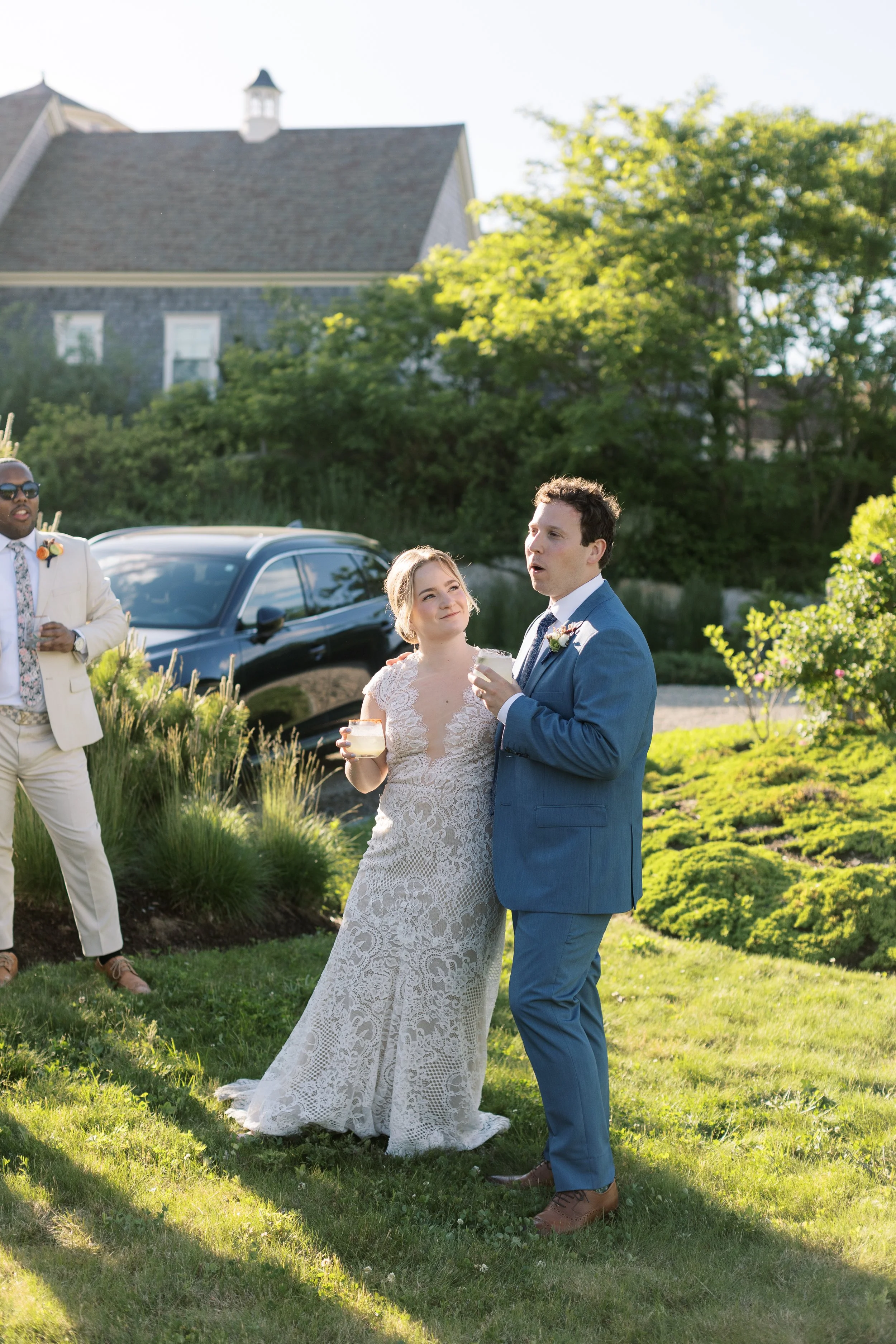 Bride and groom sharing drinks at an outdoor wedding reception, with bridesmaid and guests in the background, in a garden setting during daytime.