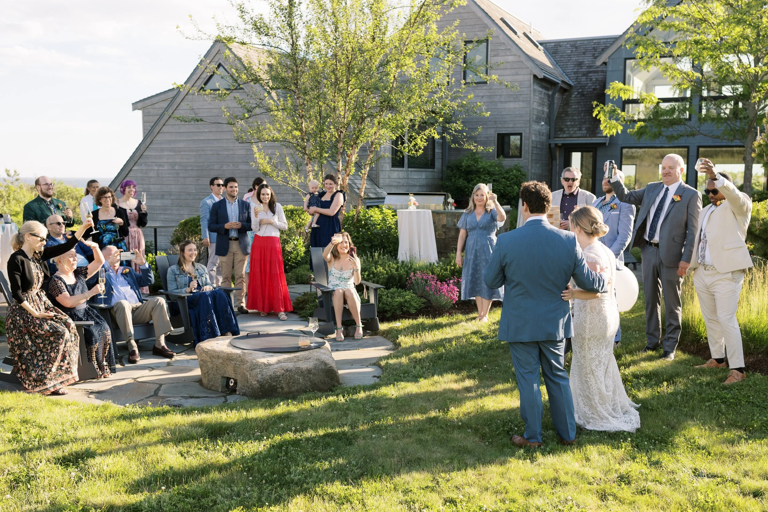 Outdoor wedding reception with guests dressed in formal attire, some taking photos or videos of the bride and groom, who are standing in a grassy area in front of a modern house, during daytime with clear weather.
