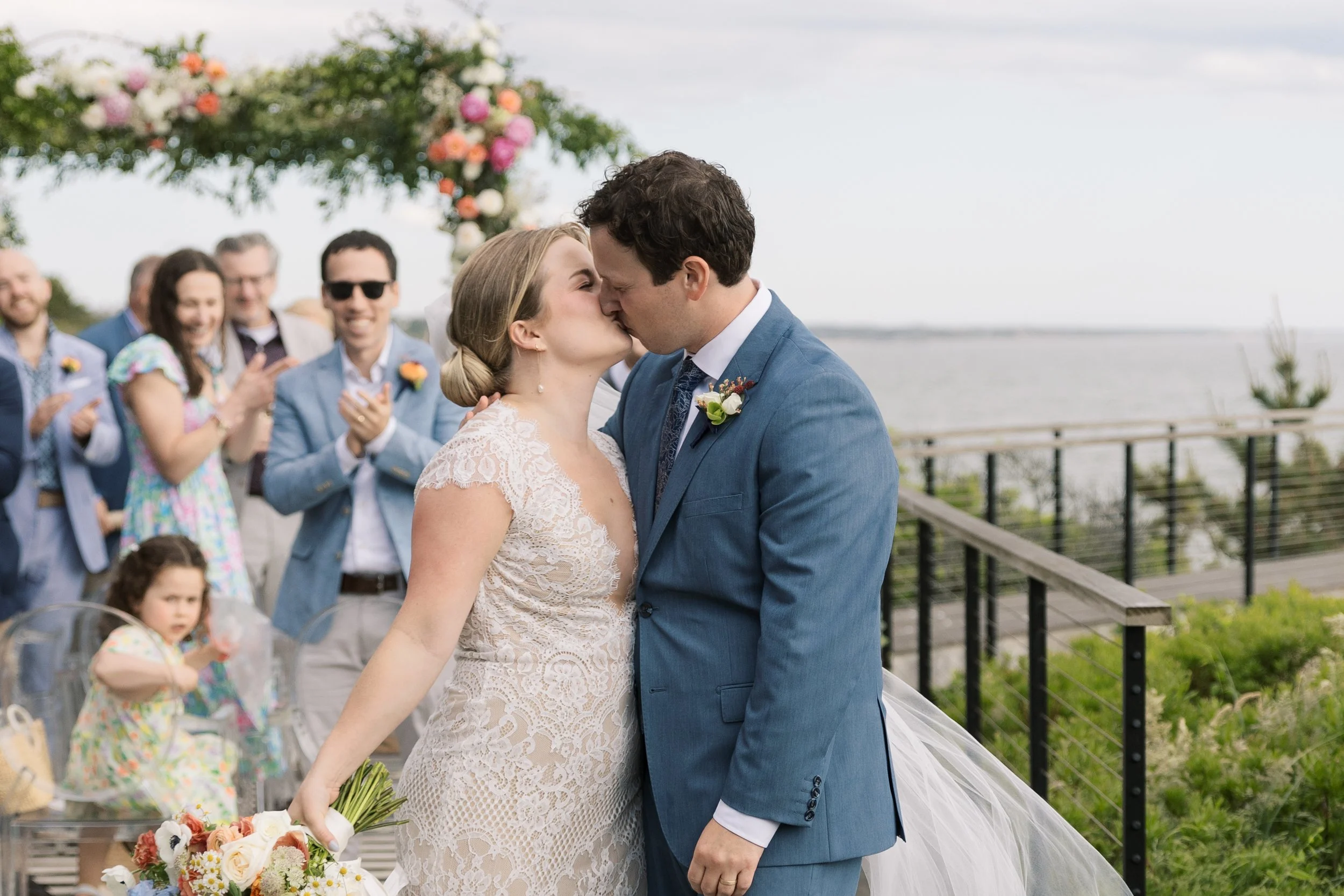 Bride and groom kissing at their outdoor wedding ceremony with guests clapping in the background, near a waterfront with a floral arch behind them.