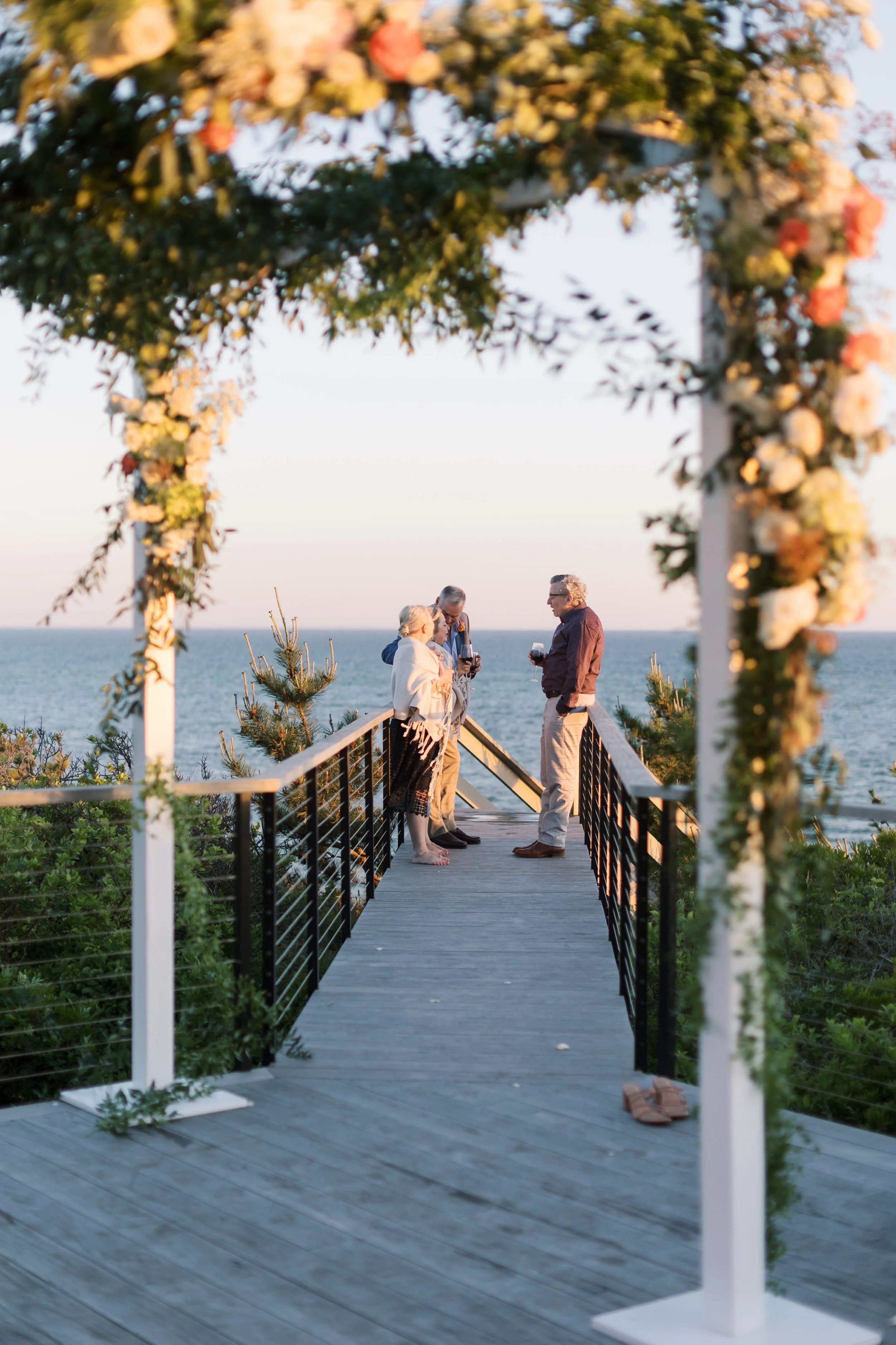 A wedding ceremony taking place outdoors on a wooden platform over the water, with four people standing under a floral arch at sunset.