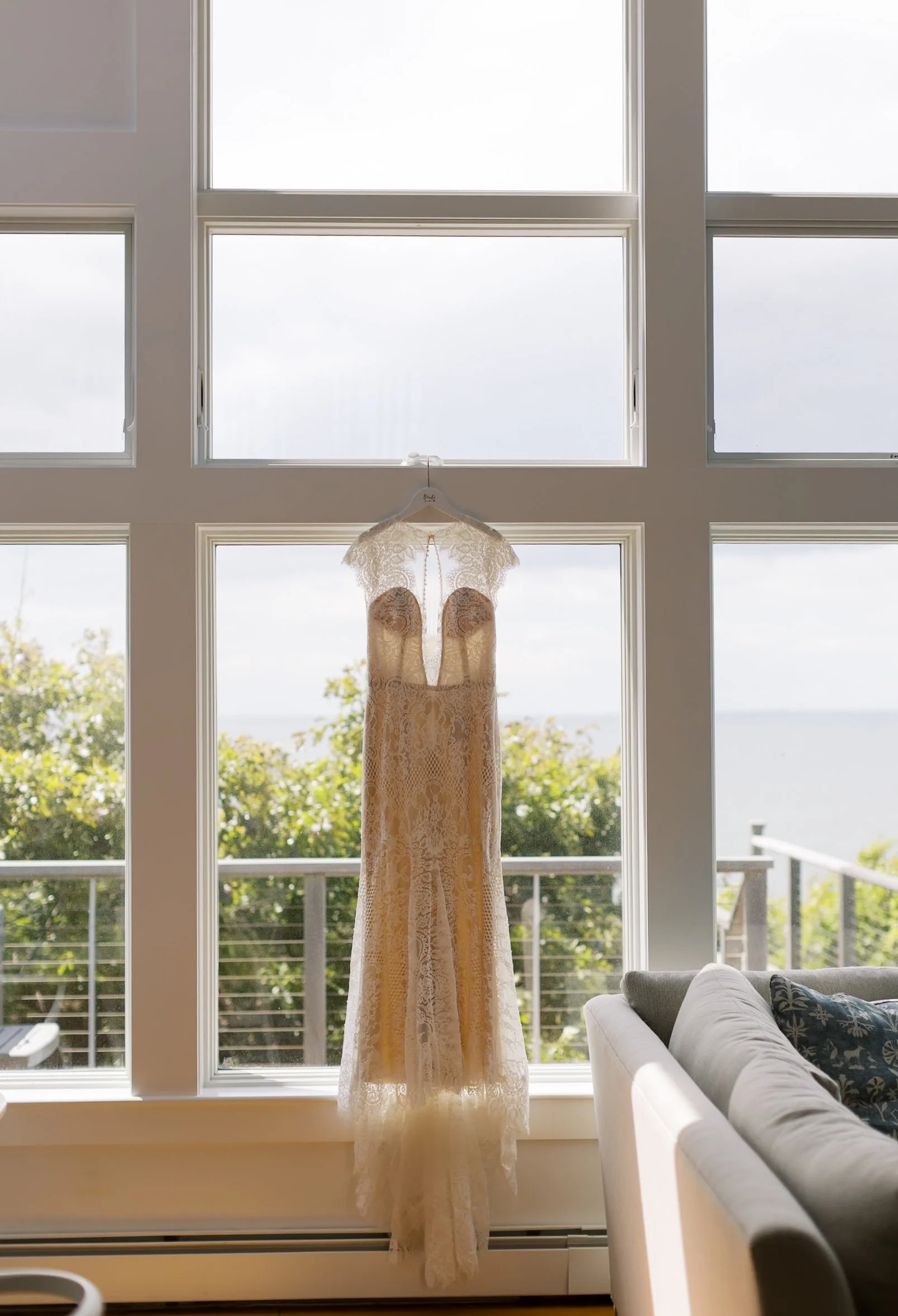 A lace wedding dress hanging on a white hanger in front of a large window with a balcony and a view of trees and water.