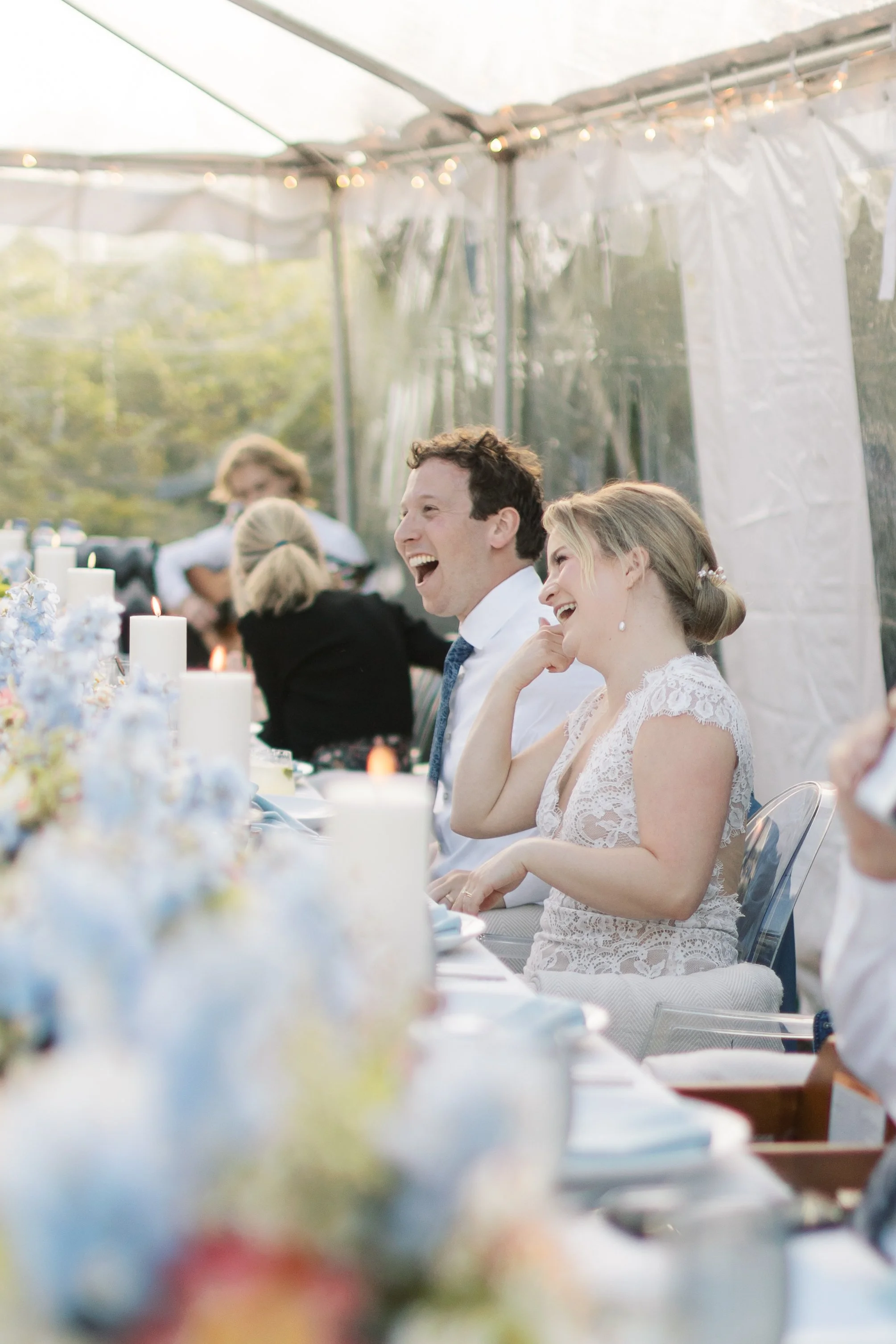 A wedding reception with a bride and groom laughing at a table decorated with candles and flowers under a clear tent.