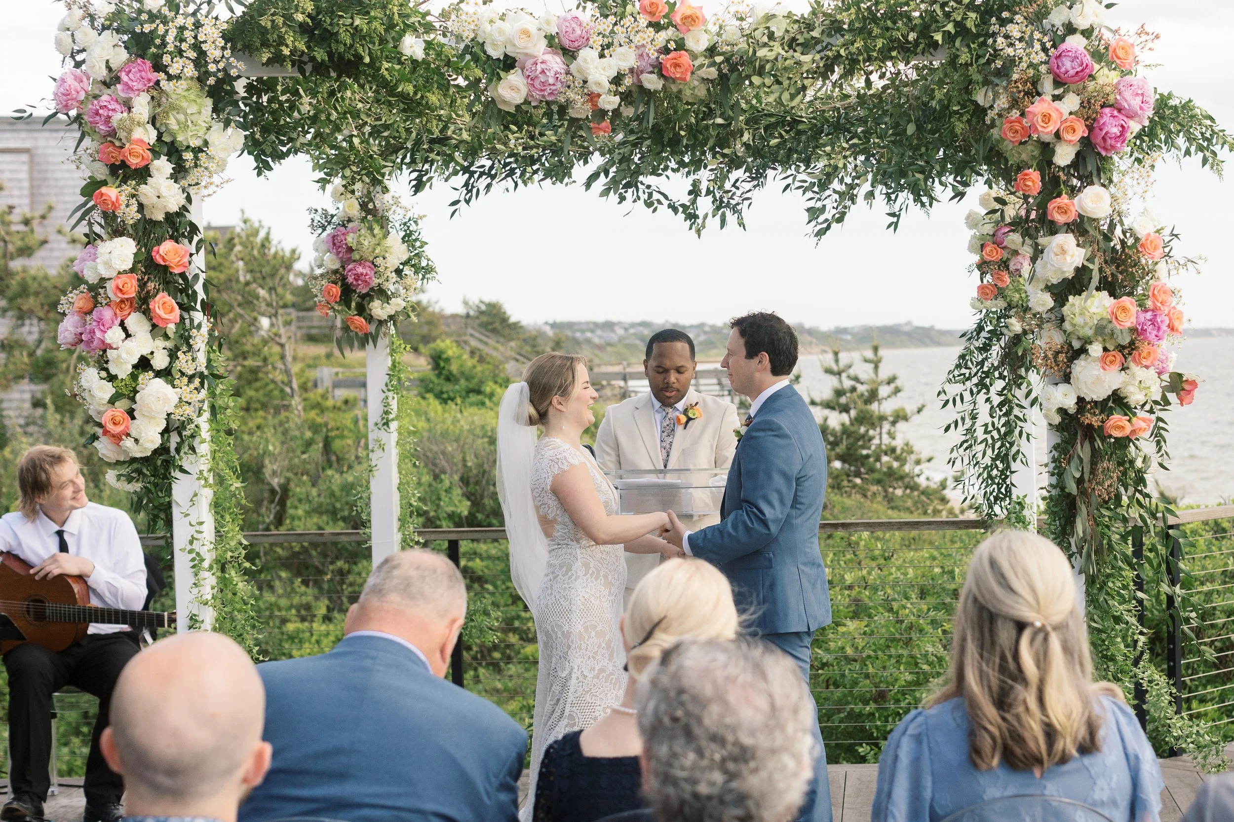 A couple gets married outdoors under a floral arch on a cloudy day, with guests seated nearby and a musician playing guitar to the left. The background shows greenery and a body of water.