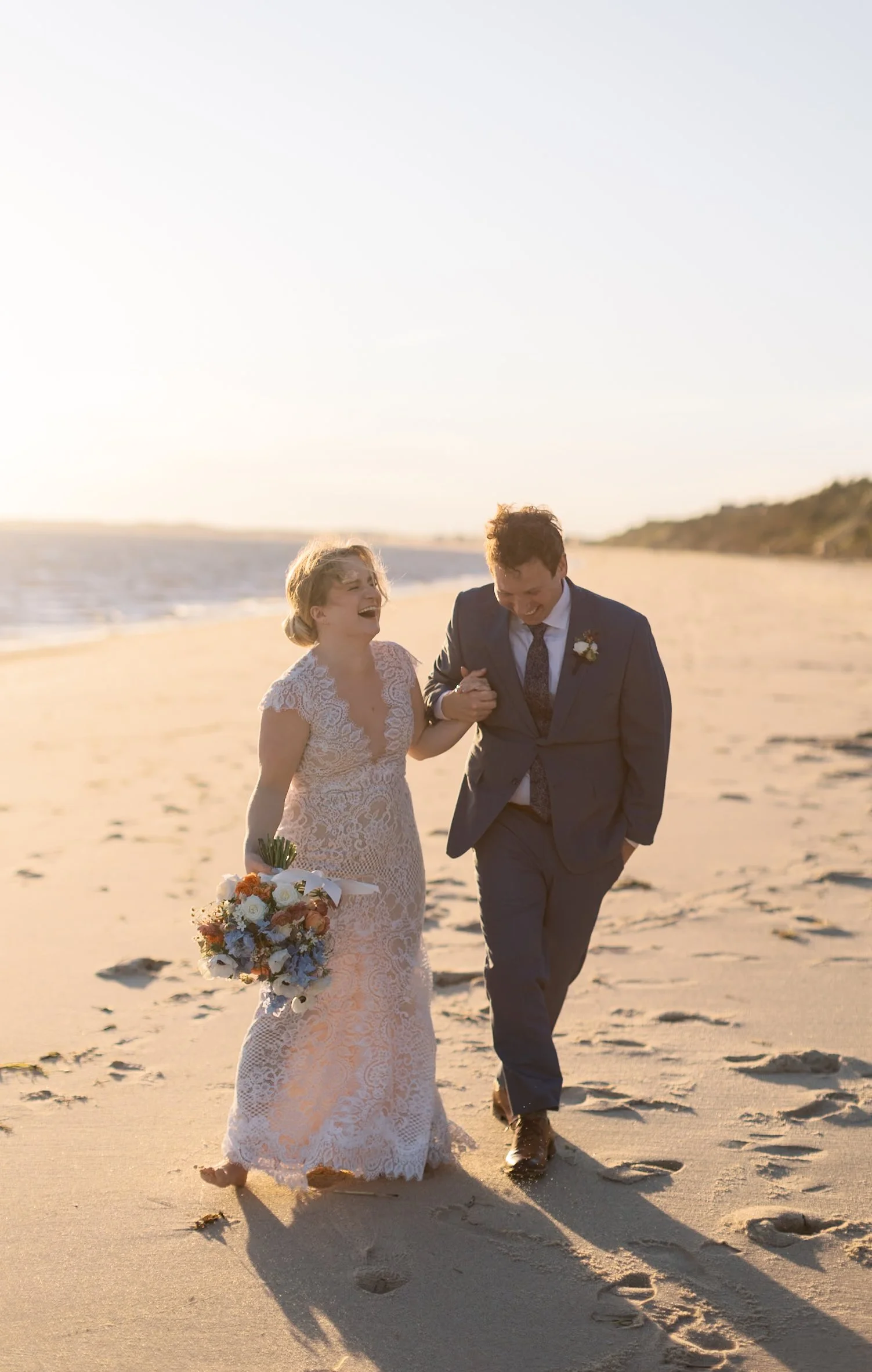 A couple, dressed in wedding attire, walking on the beach at sunset, smiling and laughing, with the bride holding a bouquet of flowers.