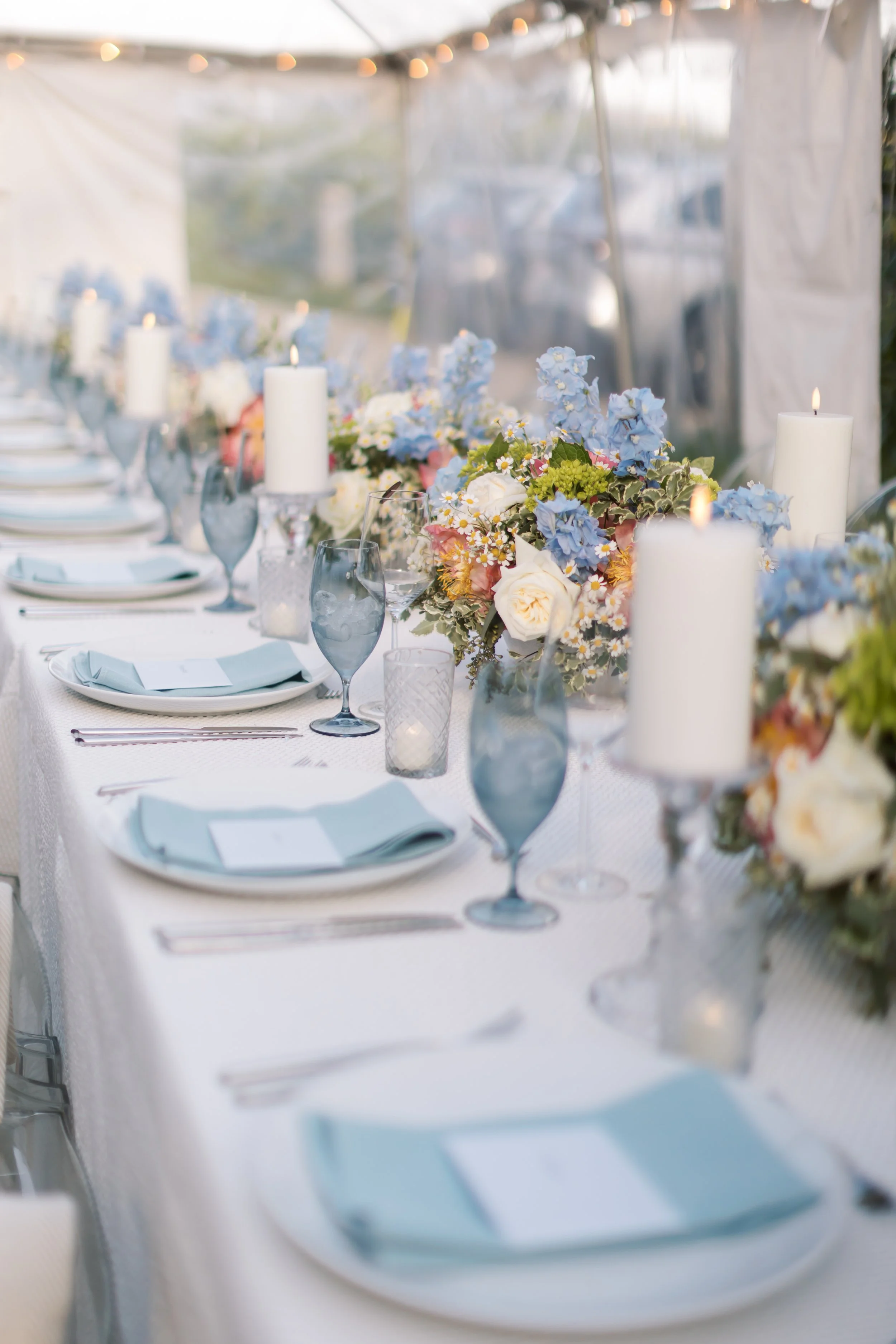 A beautifully set outdoor dining table with white tablecloth, floral centerpieces with white, pink, and blue flowers, tall white candles, blue glassware, and neatly arranged plates and silverware, under string lights tent at sunset.