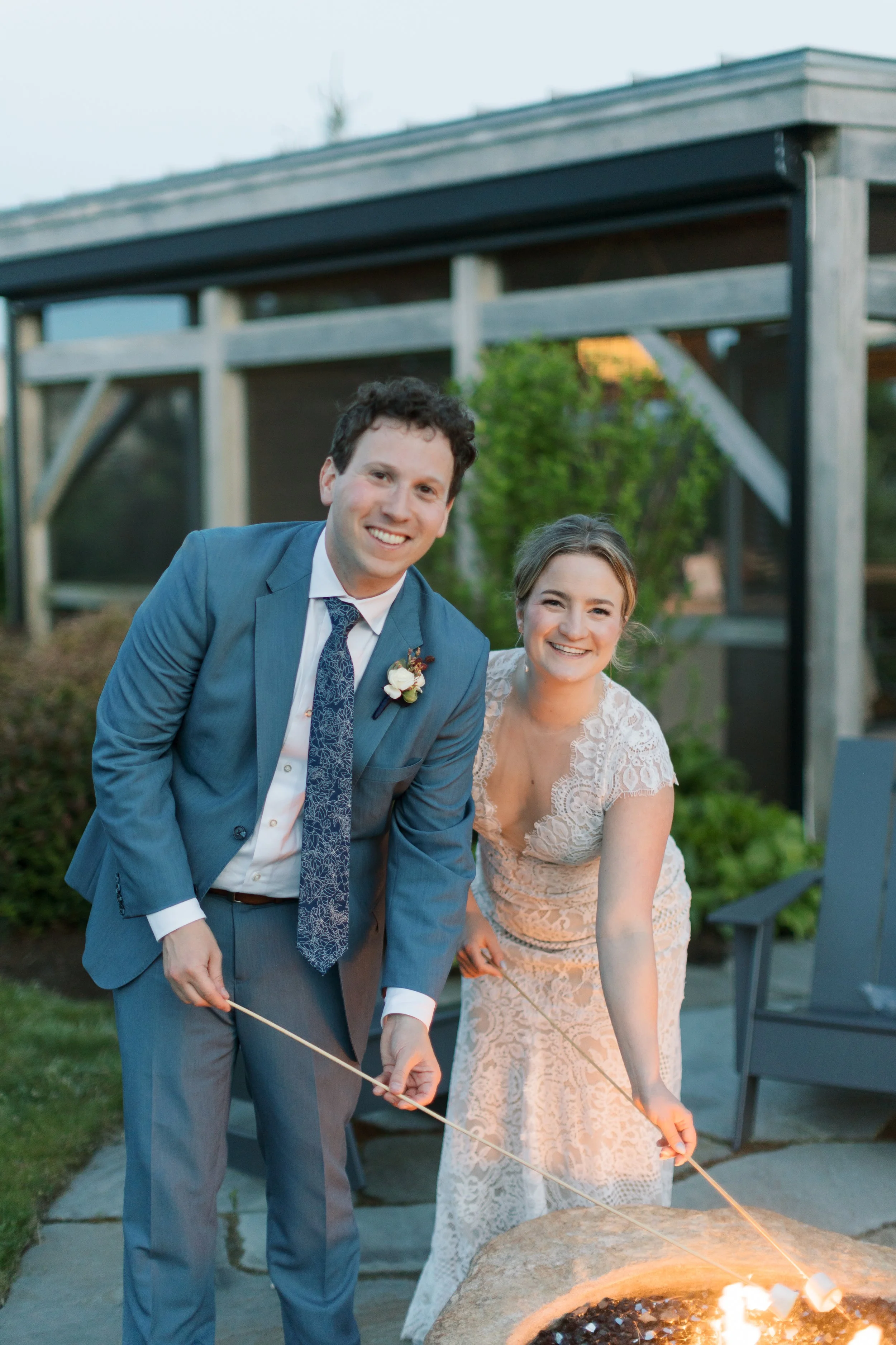 A couple in wedding attire lighting sparklers around a fire pit outdoors at sunset.