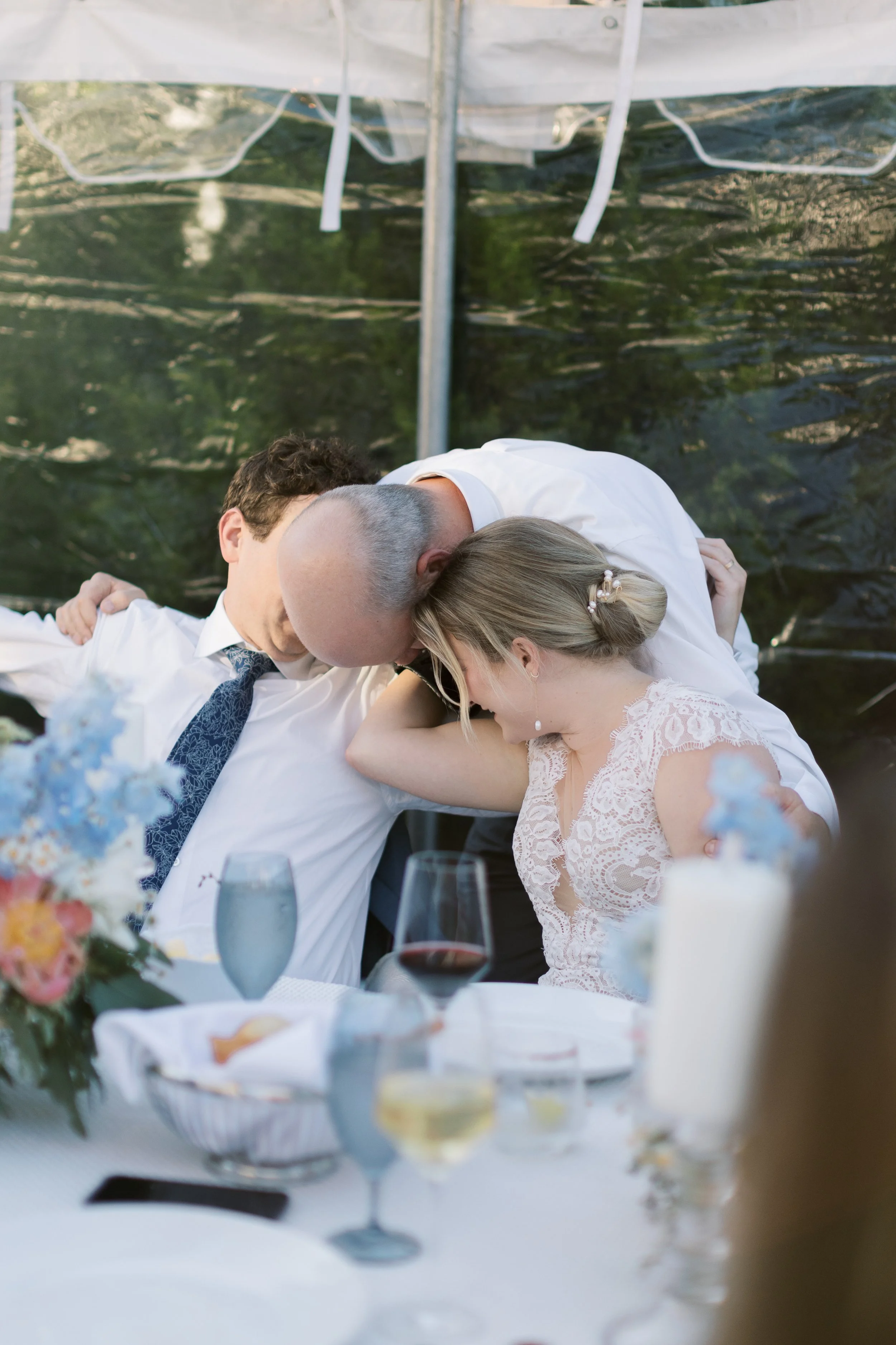 Three people sharing a tender moment at a wedding reception, with a woman in a lace dress and two men, with glasses of wine and a floral centerpiece on the table.