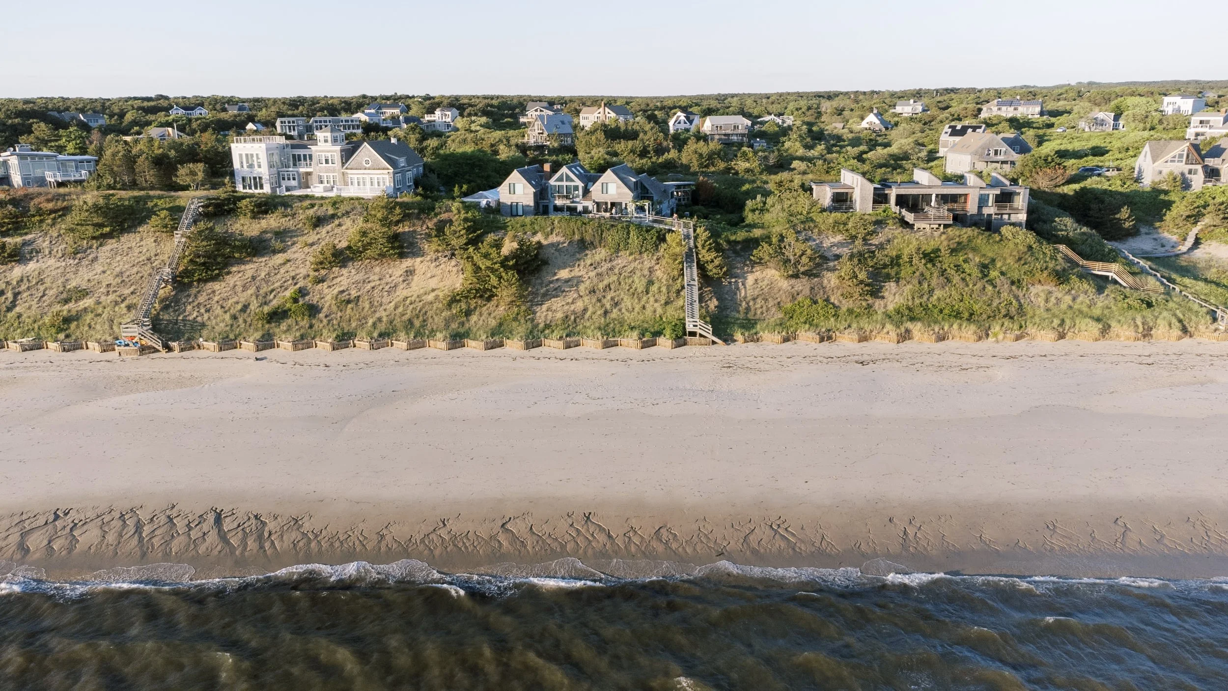 Aerial view of beach houses on dunes overlooking the ocean with sandy beach in the foreground and water at the bottom.