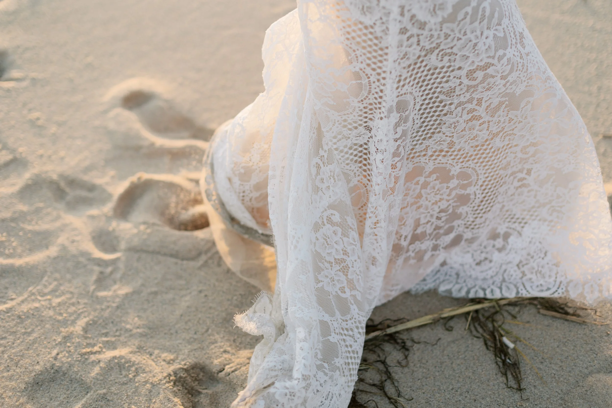 Person wearing a white lace dress sitting on sandy beach, with a piece of driftwood nearby, during sunset.