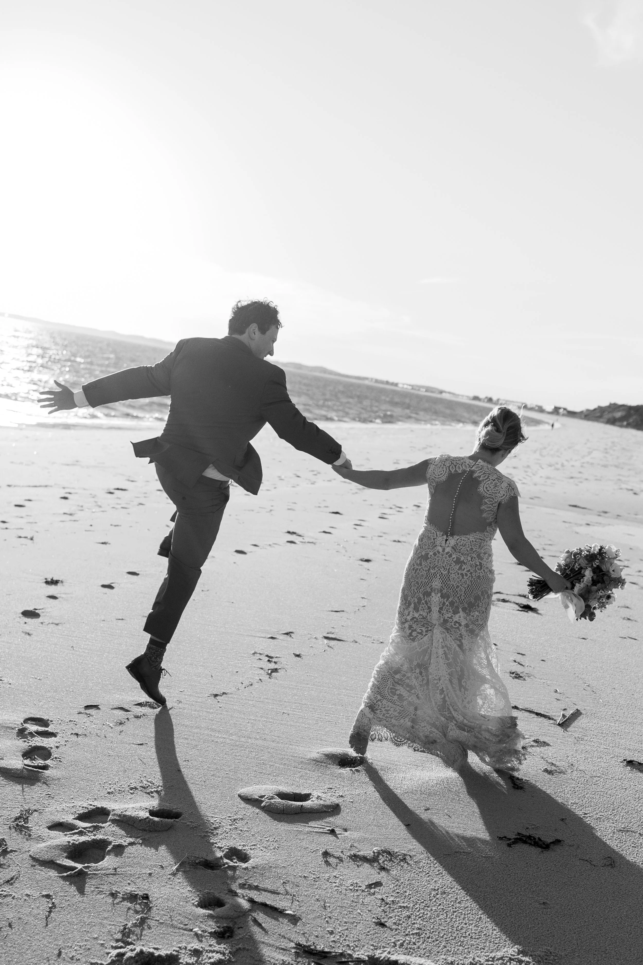 A black and white photo of a bride and groom on a beach. The groom is reaching out to hold the bride's hand as she walks barefoot carrying a bouquet. They are leaving footprints in the sand with the ocean and sky in the background.