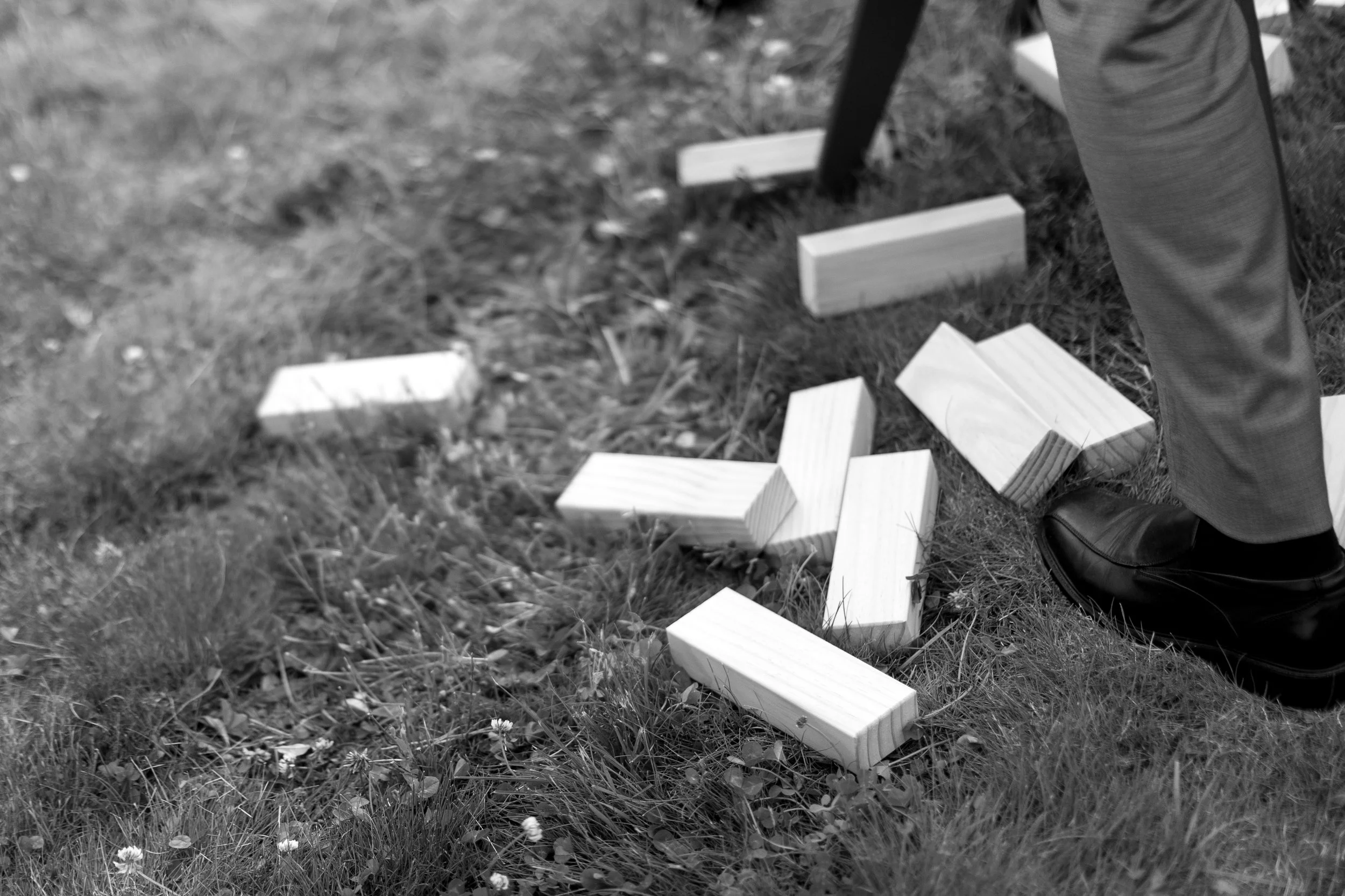 Person standing on grass ground with wooden blocks scattered around, wearing dress shoes and formal pants.