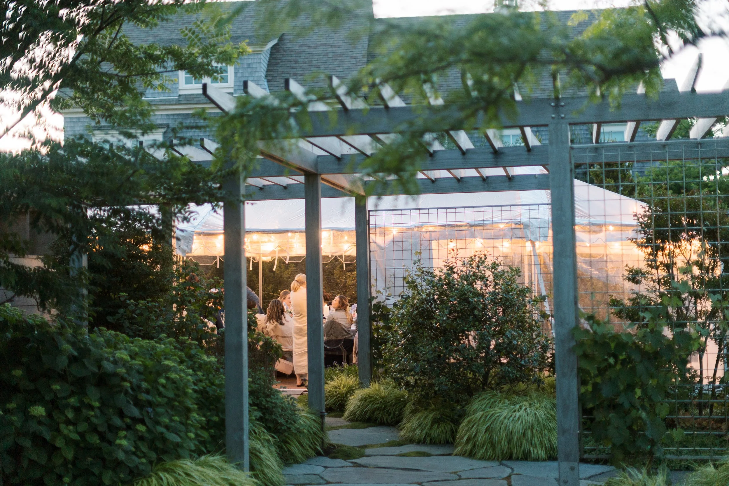 People gathered at a dinner party under a white tent, surrounded by greenery and garden structures, at dusk.