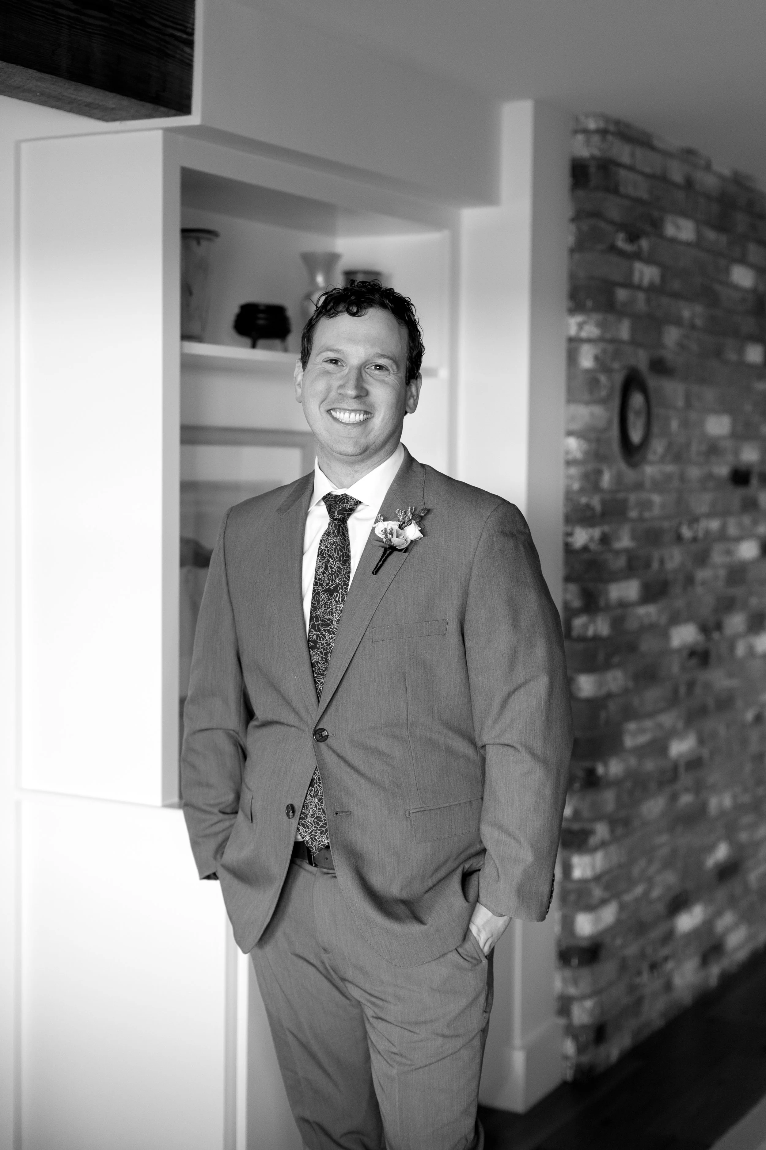 A smiling man in a suit standing indoors with his hands in his pockets, against a background of a brick wall and shelves.