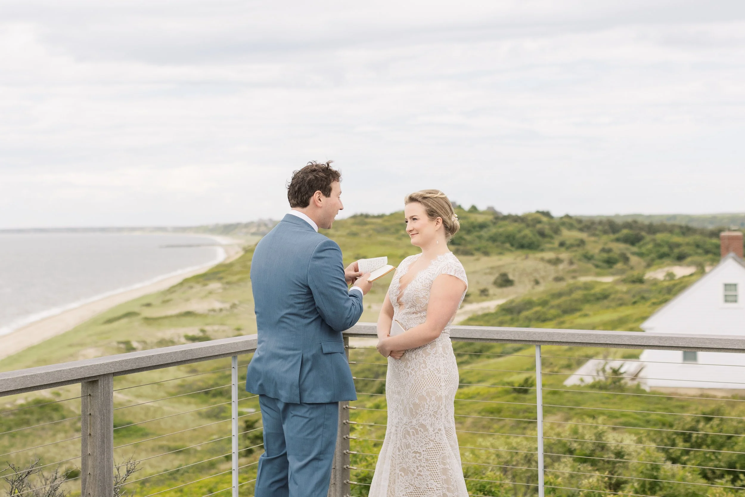 A couple getting married on a balcony overlooking a beach and green landscape, with the man reading vows from a small book and the woman smiling.