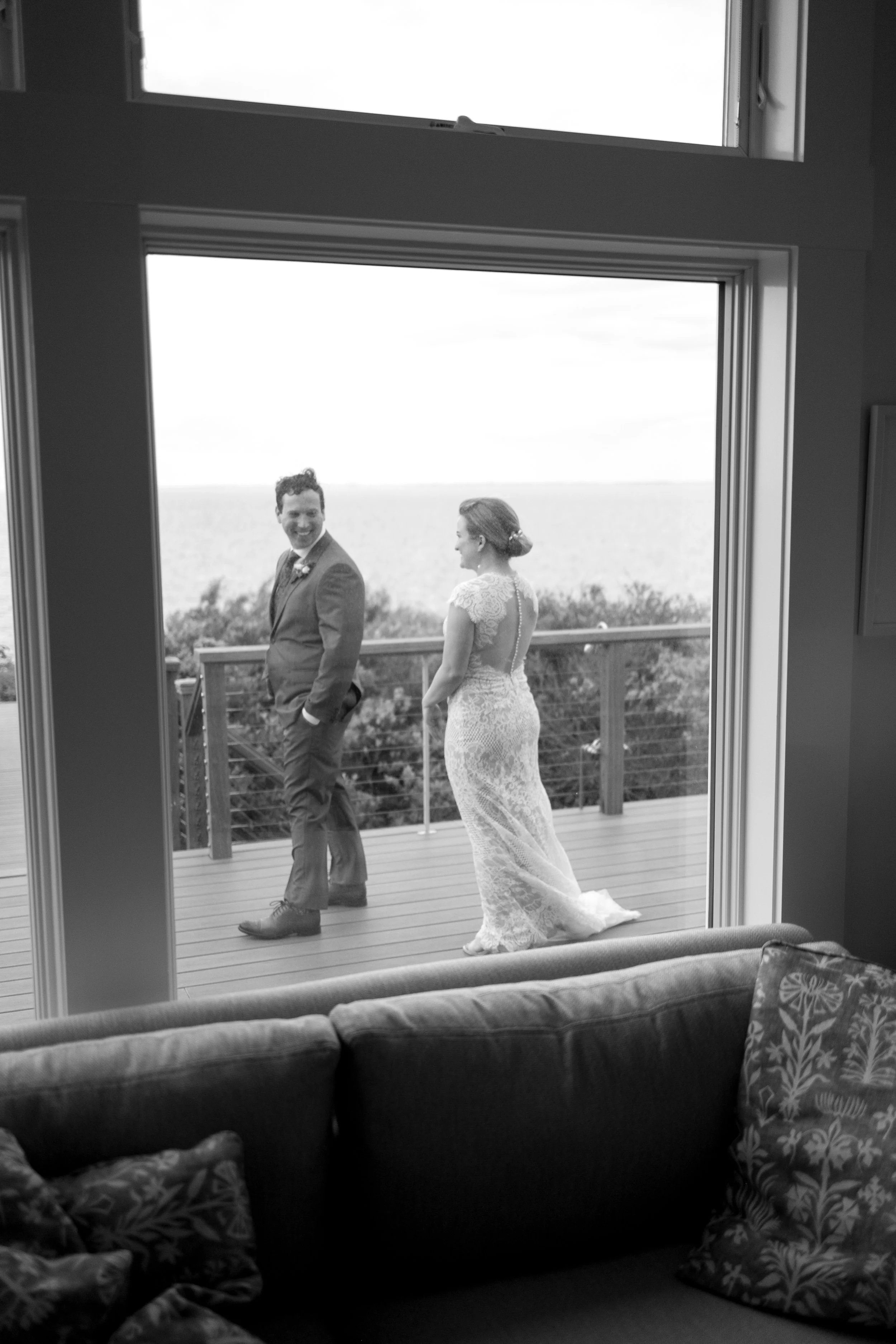 A black and white photo of a bride and groom standing on a wooden deck outside, seen through an open sliding door from inside a house. The groom is smiling and looking toward the camera, while the bride stands with her back partially turned, overlook