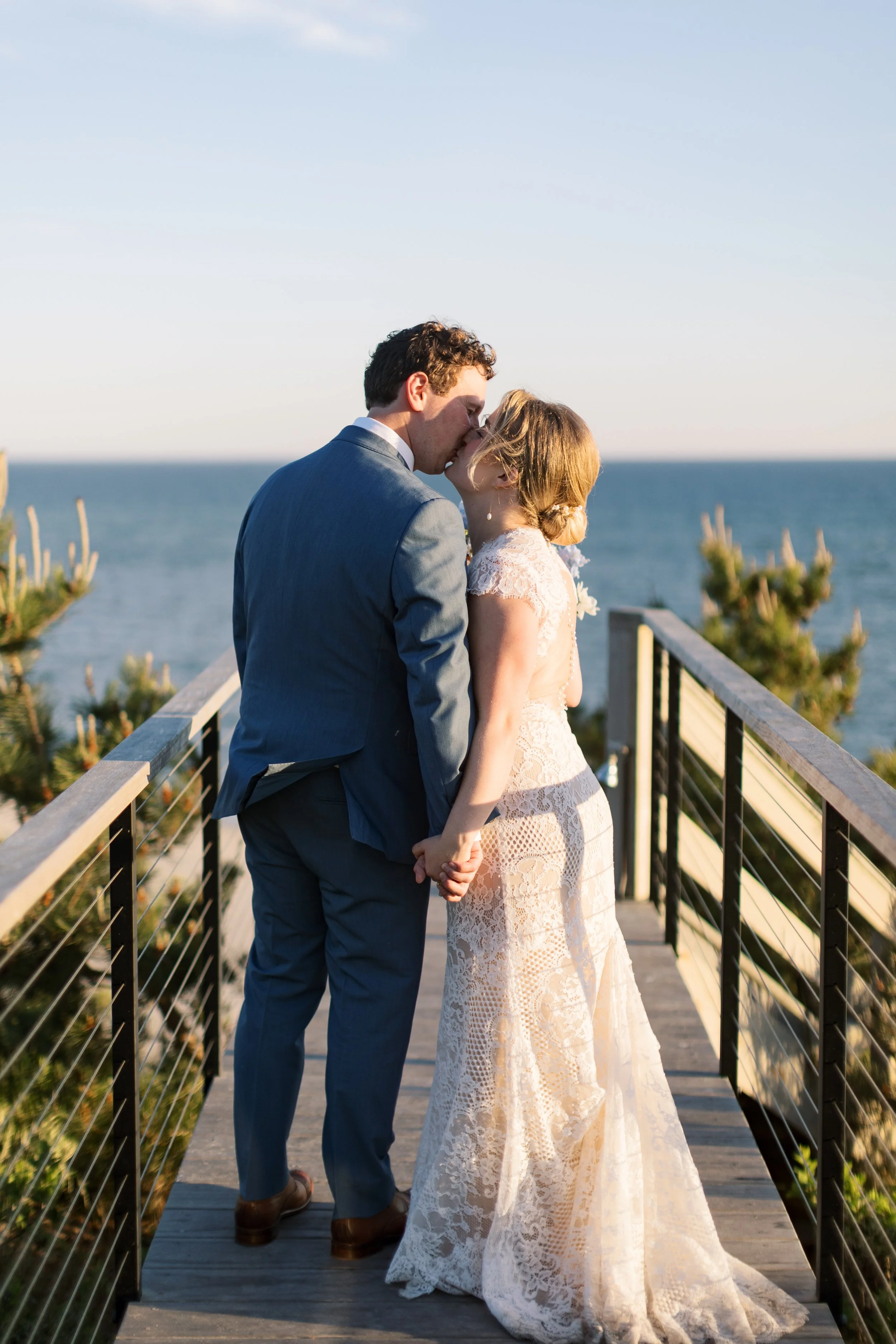 A bride and groom sharing a kiss on a wooden bridge overlooking the ocean during sunset, holding hands, with trees visible on either side.