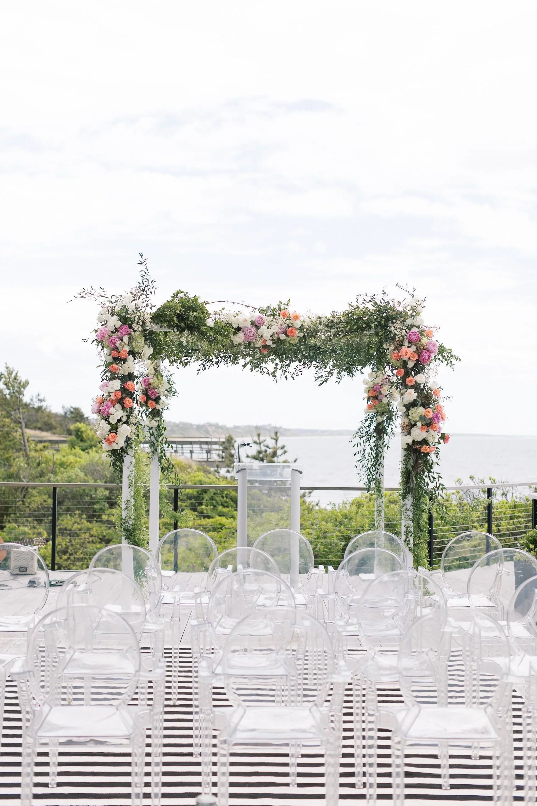 An outdoor wedding altar decorated with a floral arch, transparent chairs, and a striped black-and-white rug, overlooking a scenic body of water and greenery.