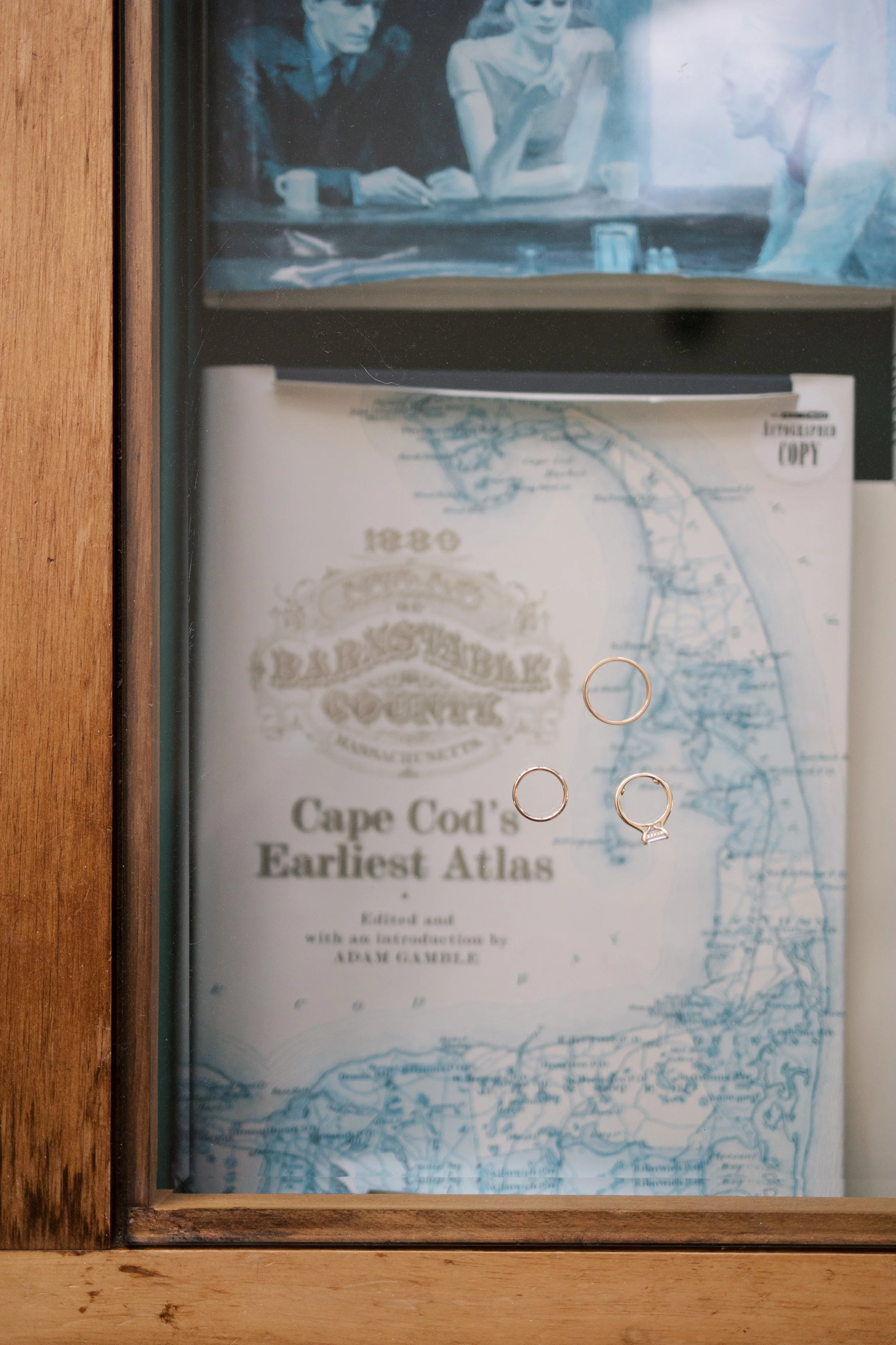 A display case with a book titled 'Cape Cod's Earliest Atlas' and three rings resting on the glass.