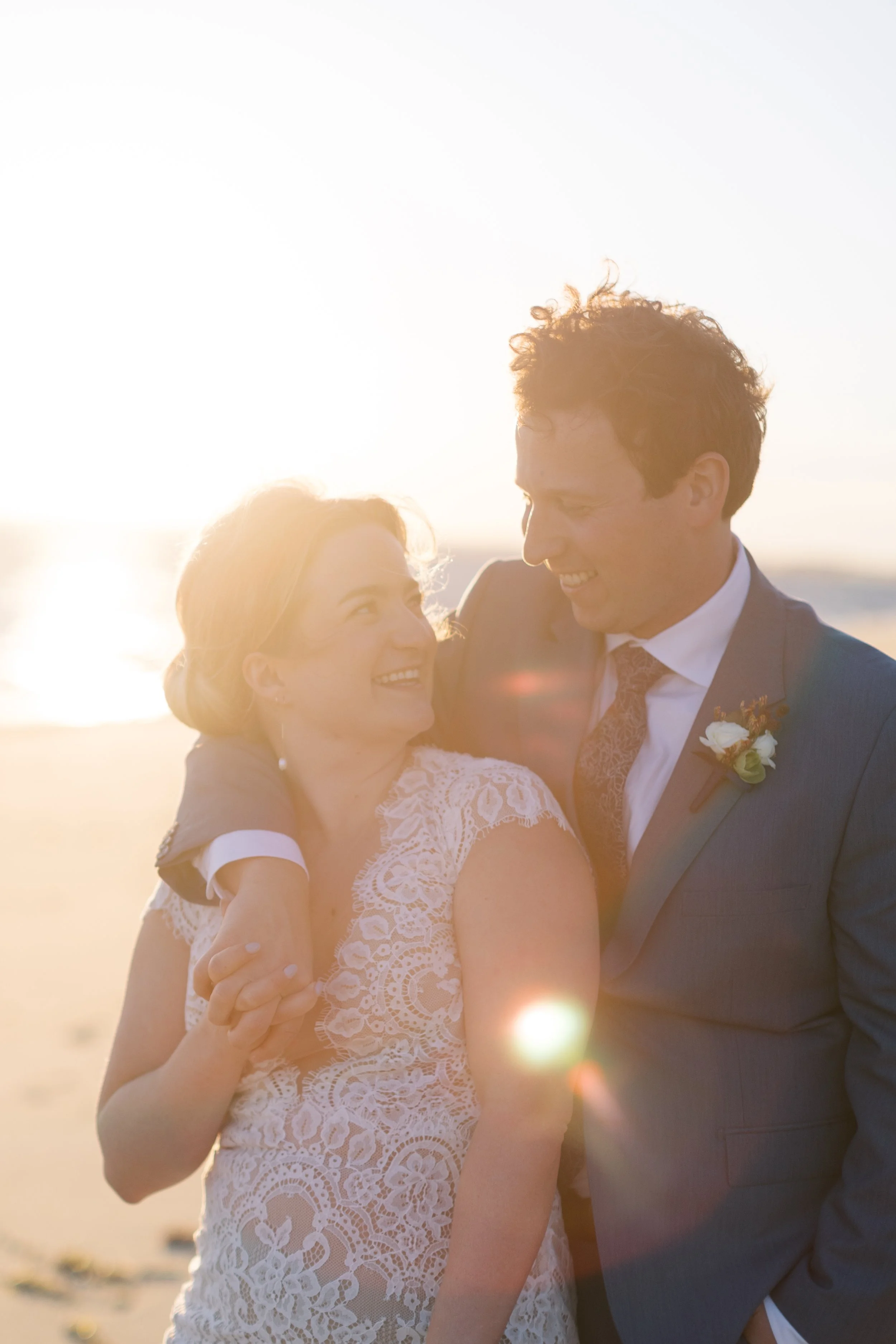 A bride and groom smiling at each other on the beach during sunset, with the sun shining brightly behind them.