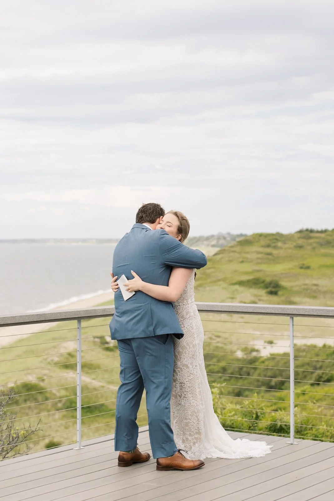 A couple embracing during their wedding on a scenic outdoor deck overlooking the ocean and green landscape.