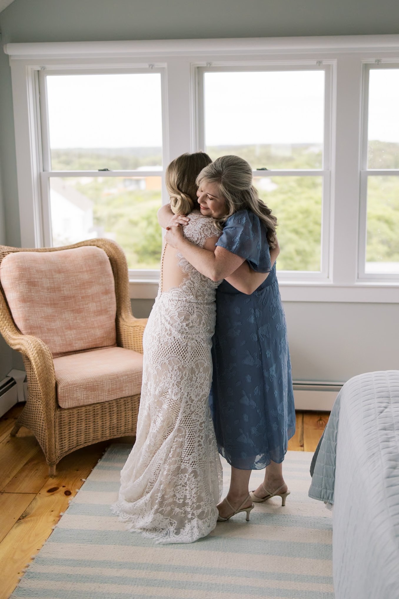 A bride in a lace wedding dress hugging a woman in a blue dress inside a bright room with large windows and a view of greenery outside.
