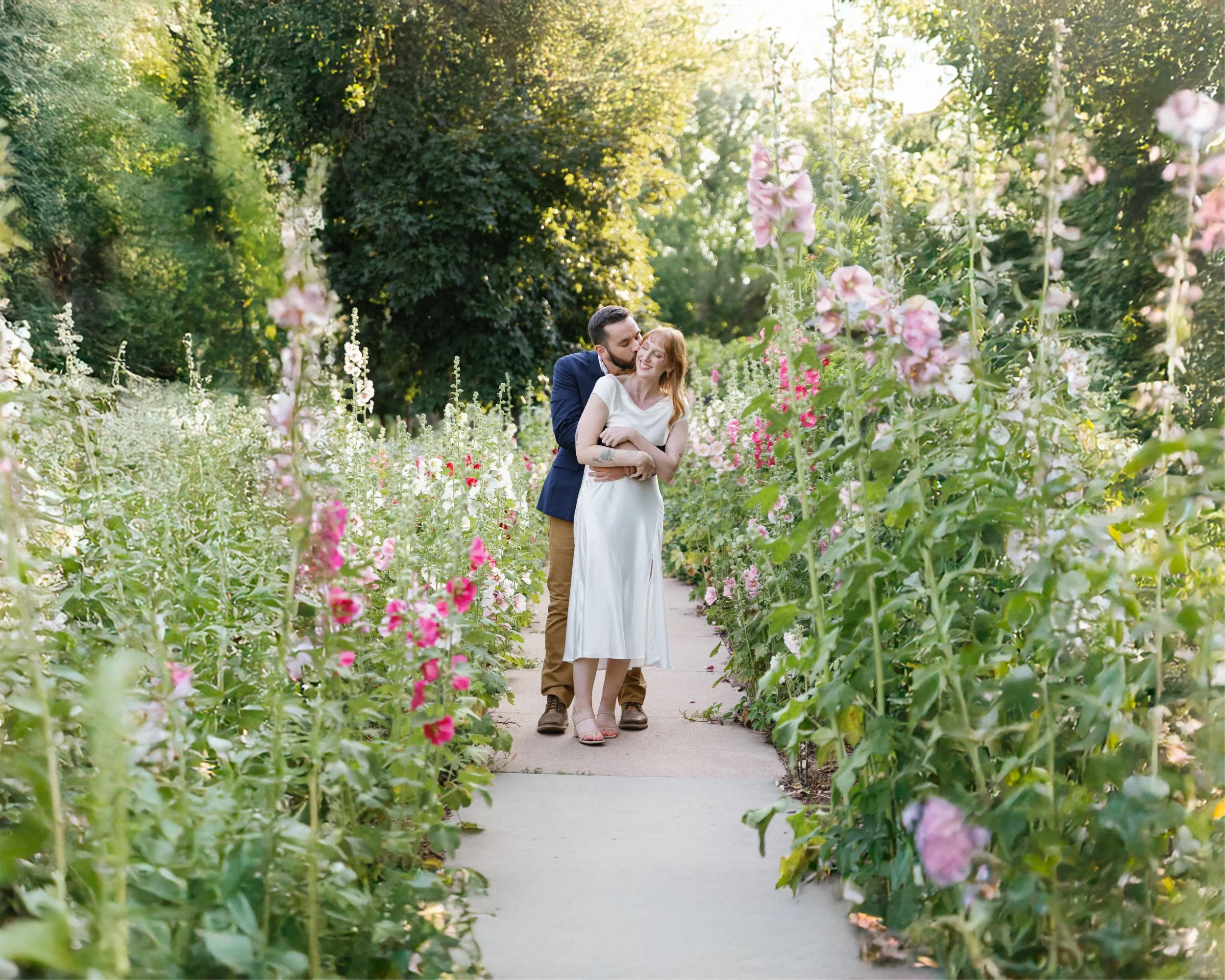 A couple embracing and kissing in a garden with tall flowering plants and trees, sunlight filtering through the leaves.
