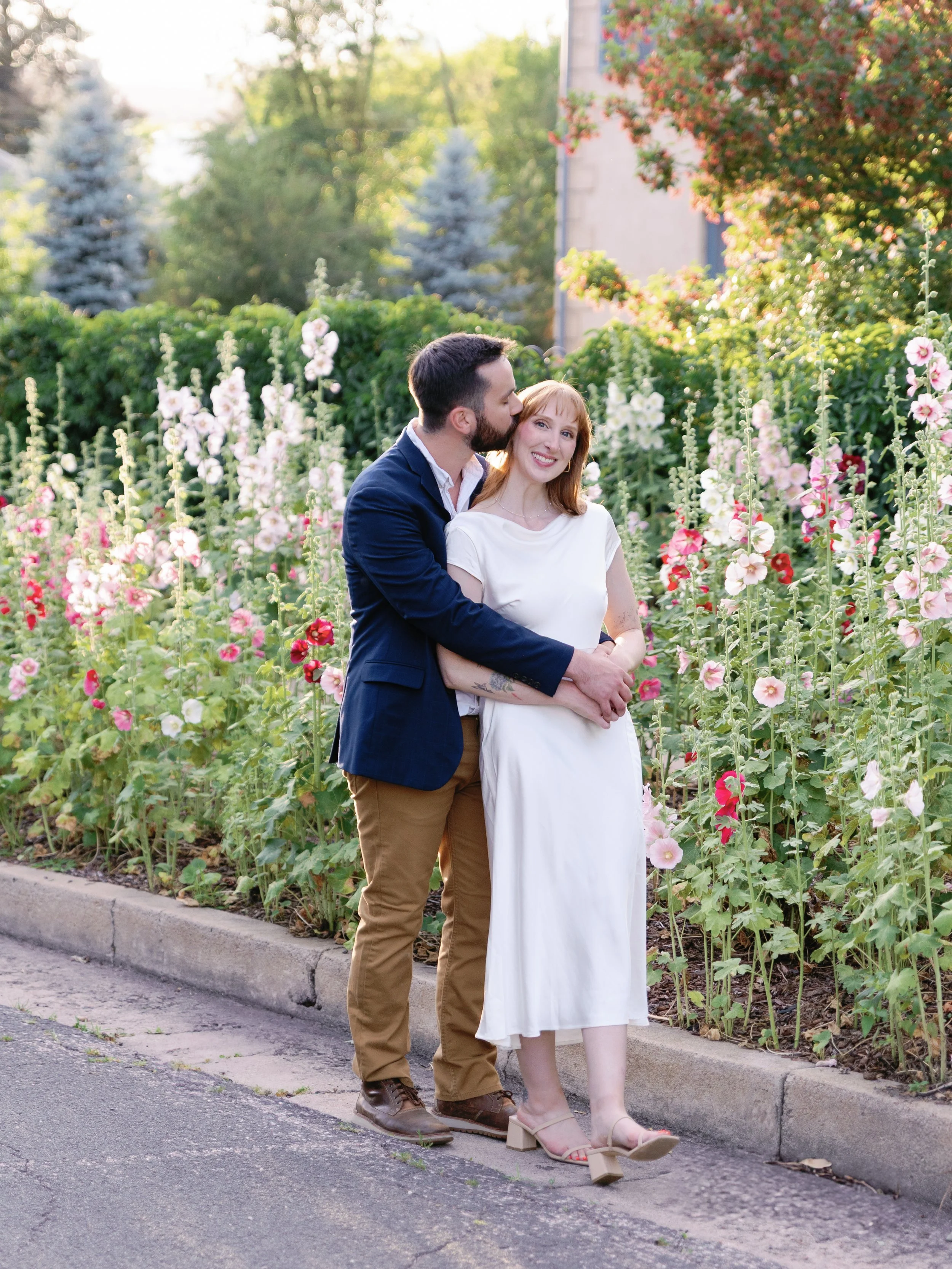 A couple standing on a sidewalk next to a flower bed with pink and white hollyhocks. The man is kissing the woman's temple, and she is smiling. The woman has red hair and is wearing a white dress with beige heels, while the man has dark hair and is w
