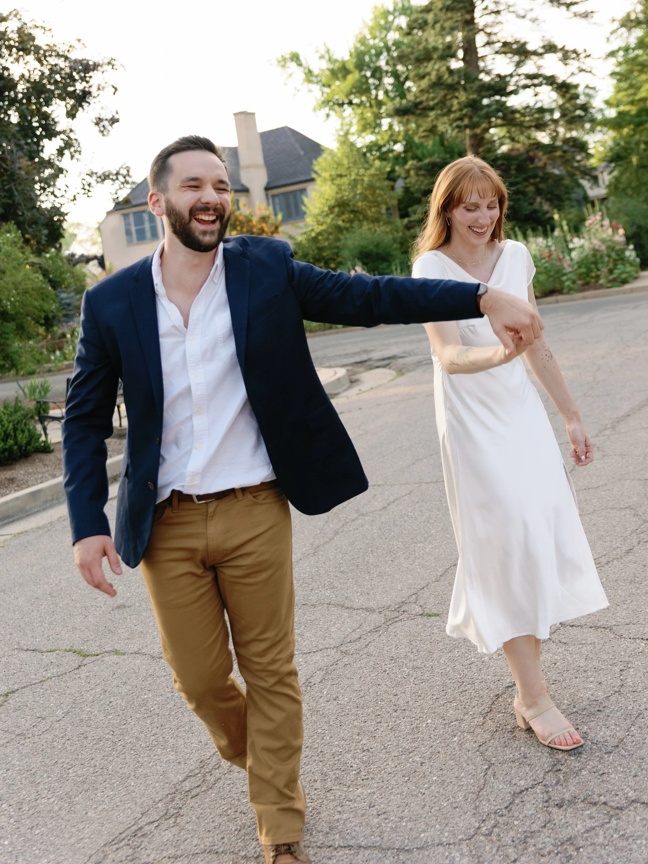 A smiling man and woman walking hand in hand outdoors, with the man leading. The man is wearing a navy blazer, white shirt, and tan pants, and the woman is in a white dress and beige heels. They are on a paved street with greenery and a house in the 