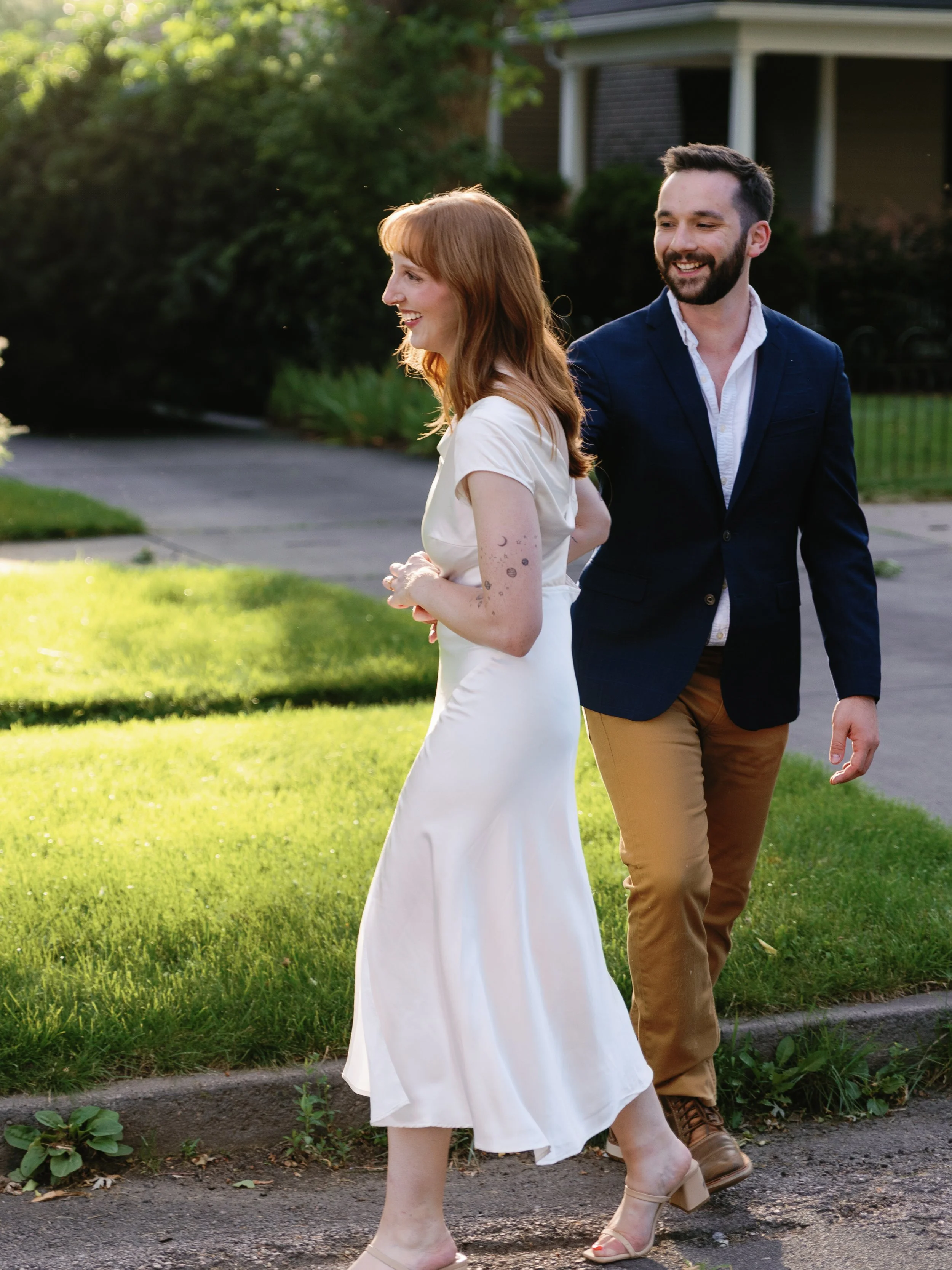 Two people, a woman with red hair in a white dress and a man with dark hair and a beard in a navy blazer, are walking outdoors on a sunny day, smiling and talking.